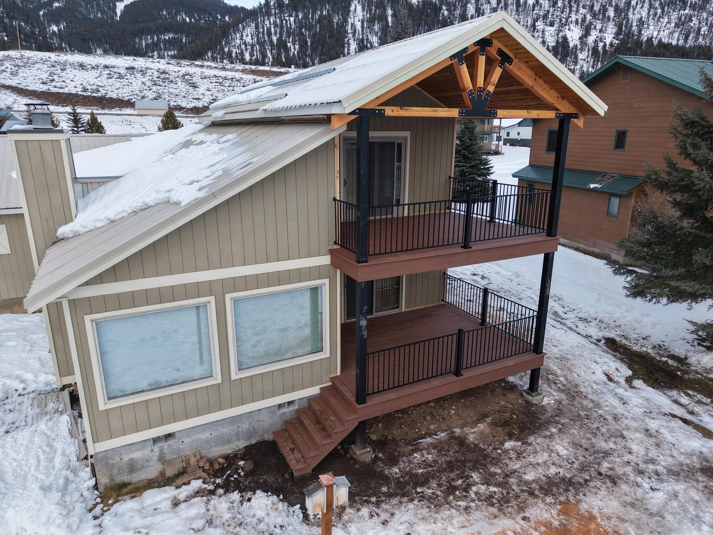Two-story house with a newly constructed balcony and stairs outside, snow on the roof and ground, with a mountain and trees in the background.