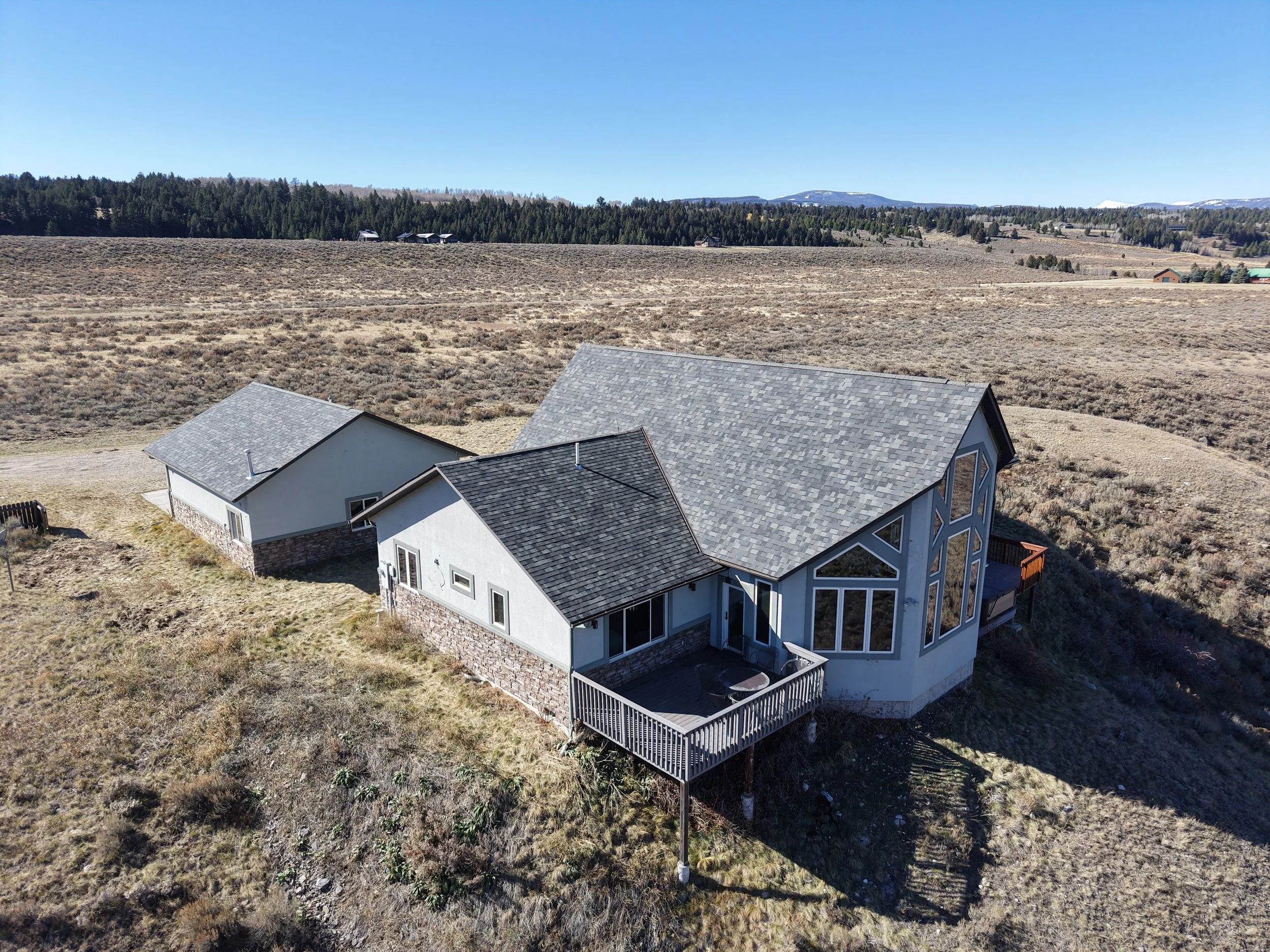 An aerial view of a house with a large front-facing window, a deck, and a barn in a vast open field with dry grass, distant trees, and mountains under a clear blue sky.