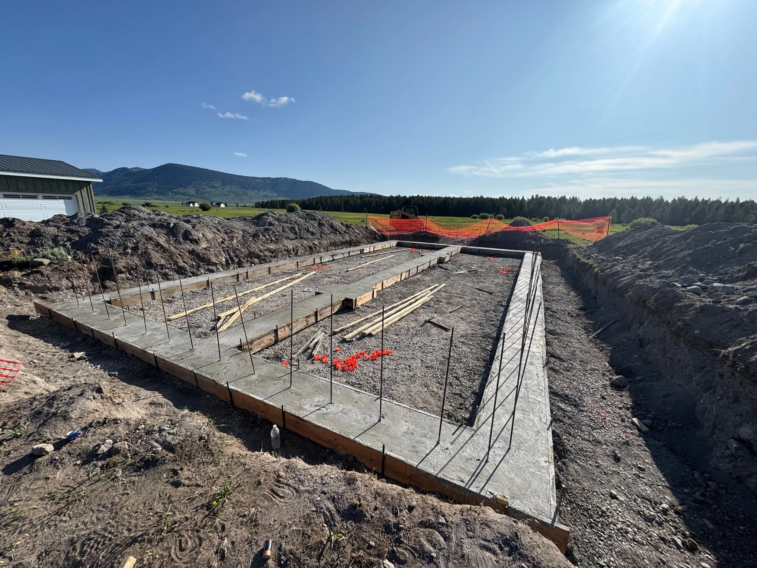 Construction site with a concrete foundation, rebar, and wooden formwork in a rural area with mountains and green fields in the background under a clear blue sky.