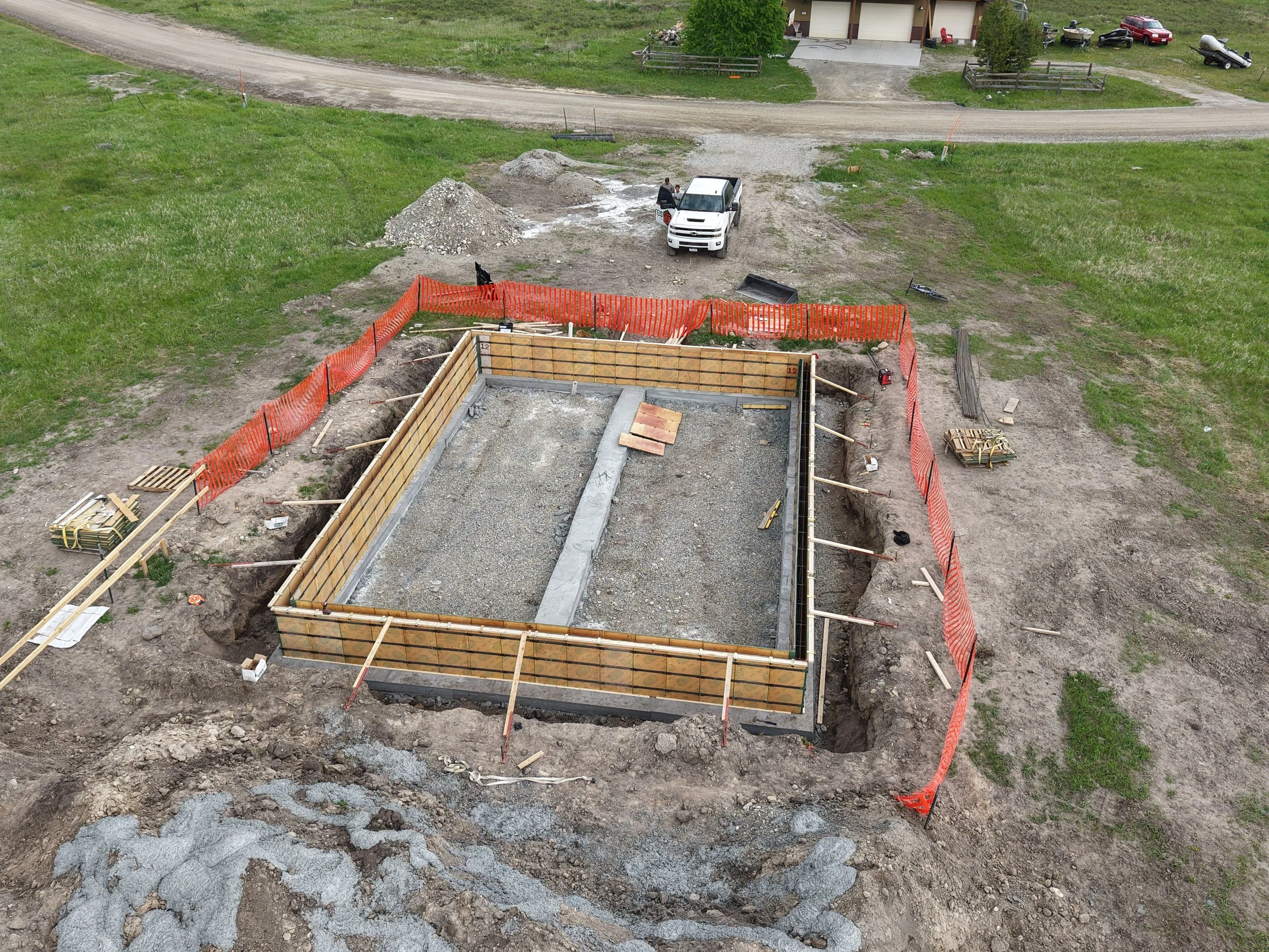 Construction site with a foundation in progress, surrounded by orange safety fencing, with construction tools and materials nearby, and a white truck parked at the site. A house and a few vehicles are visible in the background.