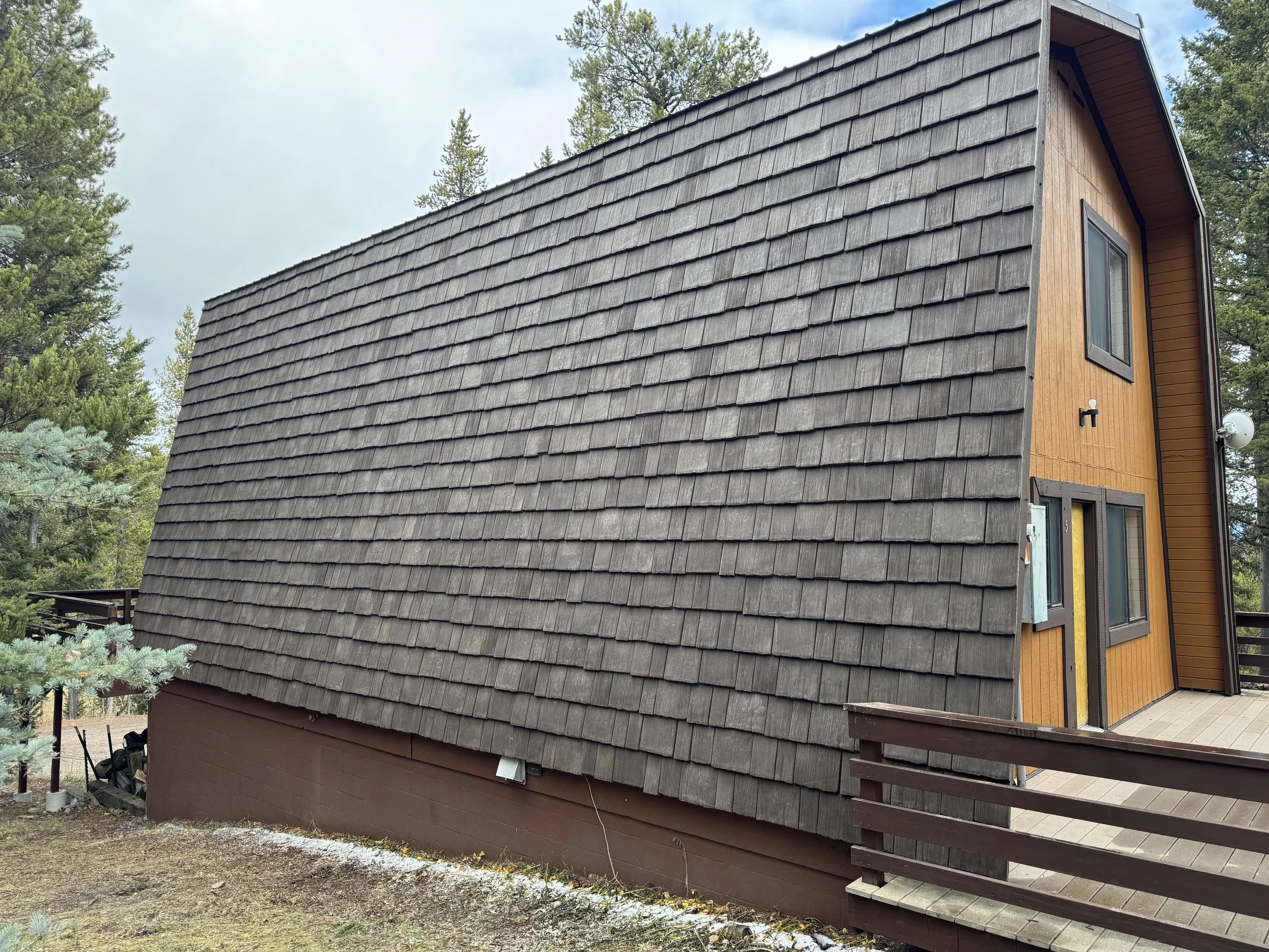 Small house with a steep roof made of weathered wooden shingles, wooden siding on the side, and a deck with a railing.