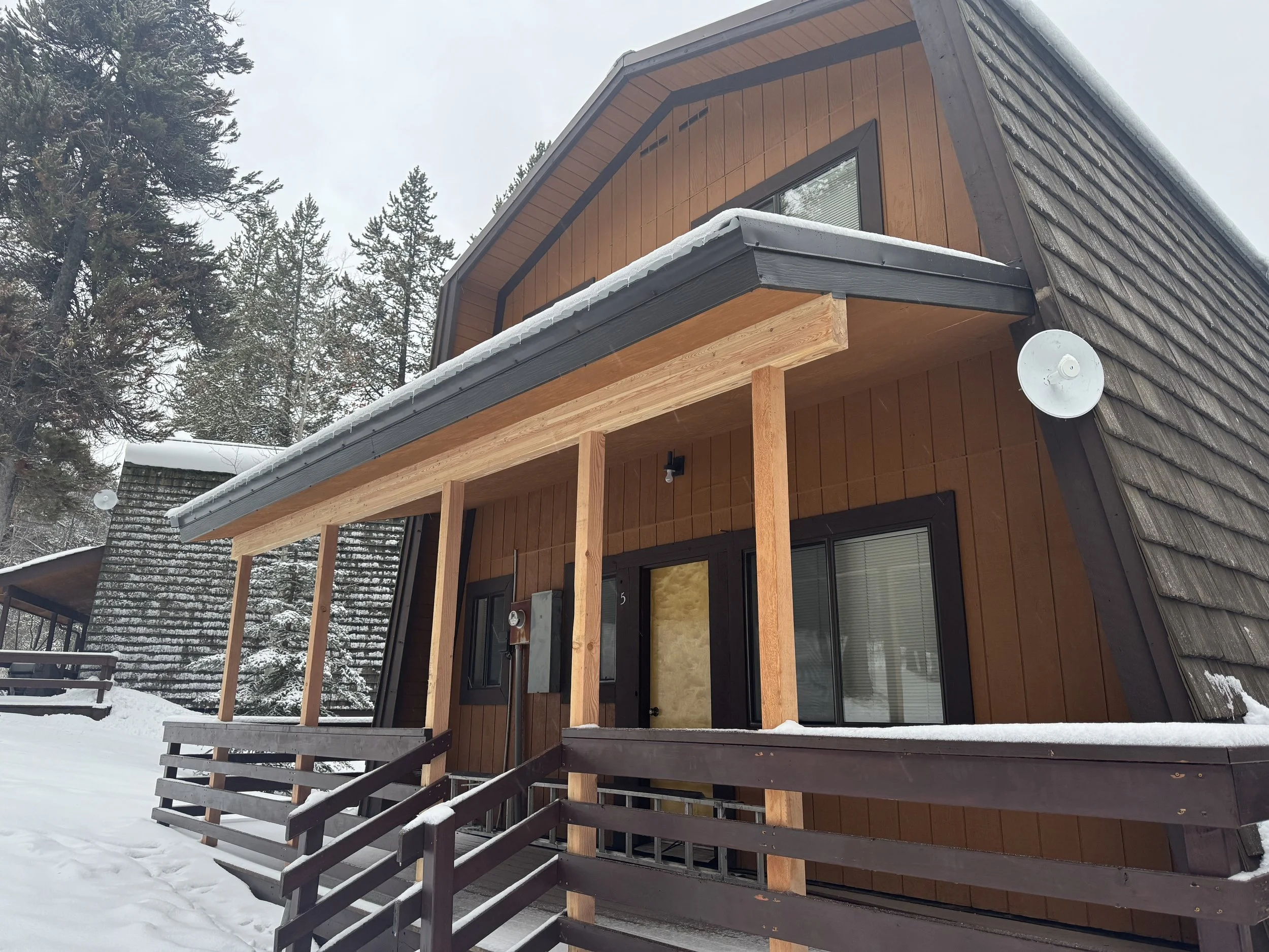 A modern wooden house with a snow-covered outdoor porch, surrounded by snow and tall pine trees, during winter.