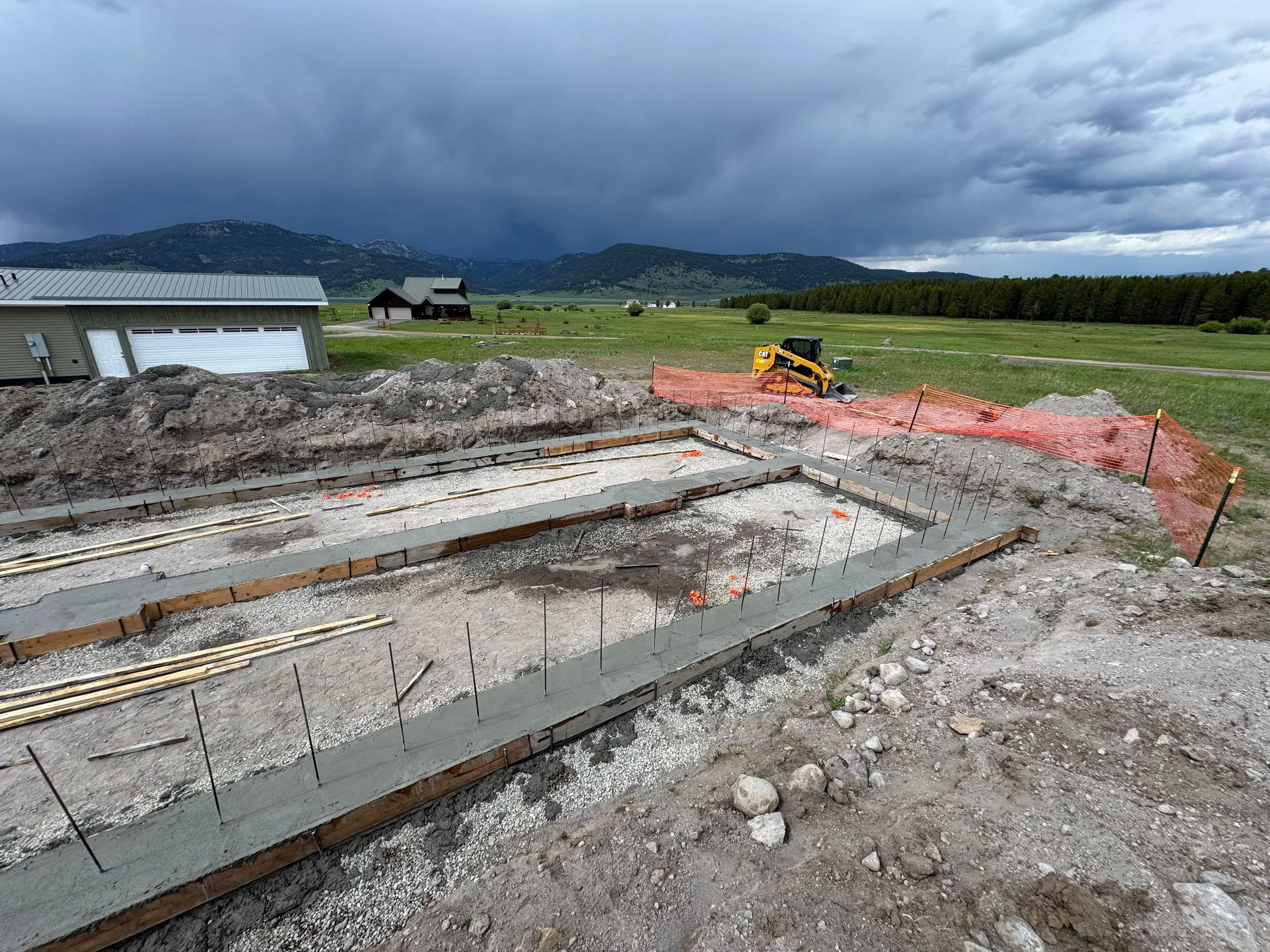Construction site in a rural area with a foundation in progress, surrounded by green fields and mountains under a cloudy sky.