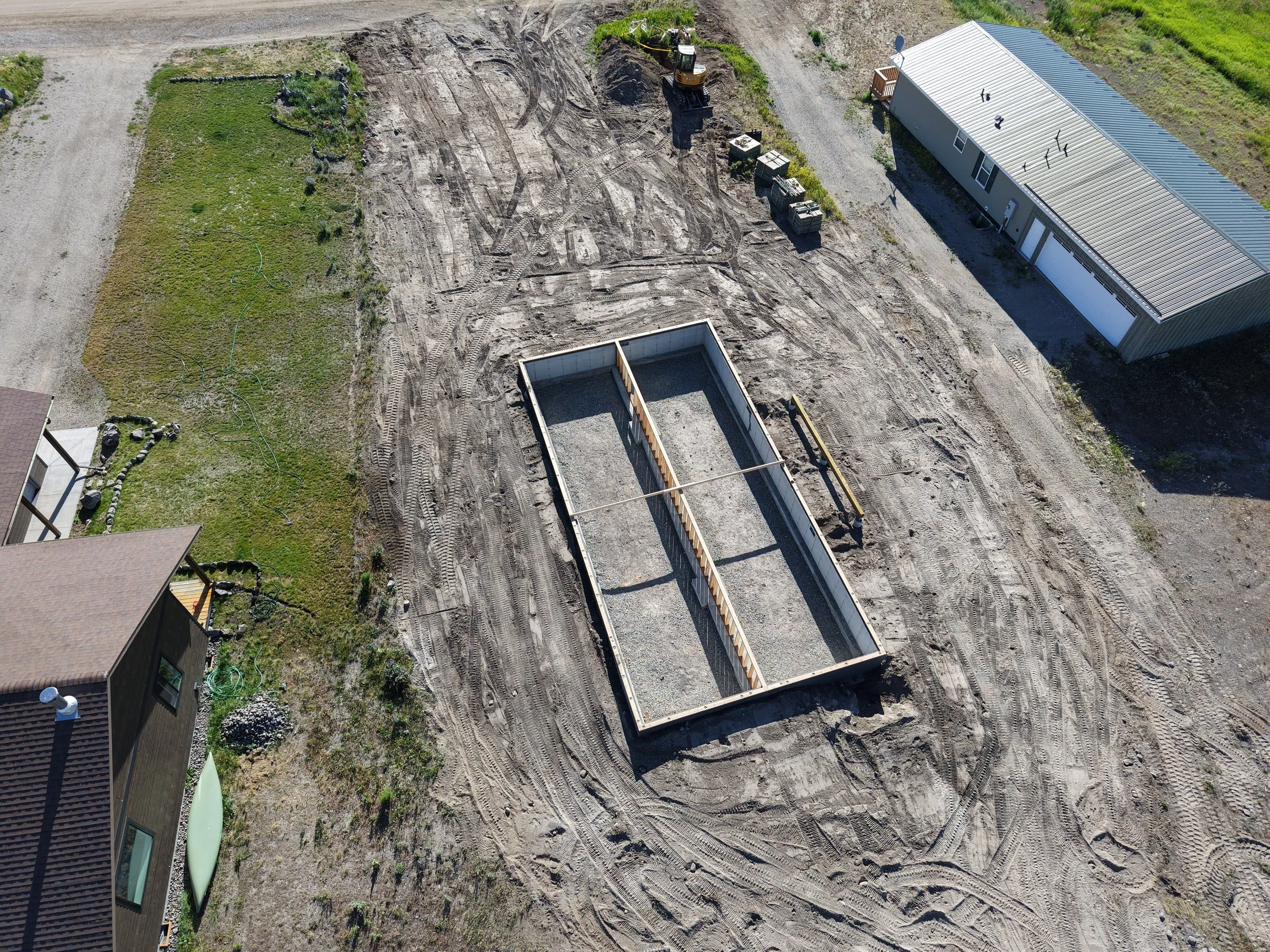 Aerial view of a residential construction site showing a foundation outline, construction equipment, and neighboring houses.