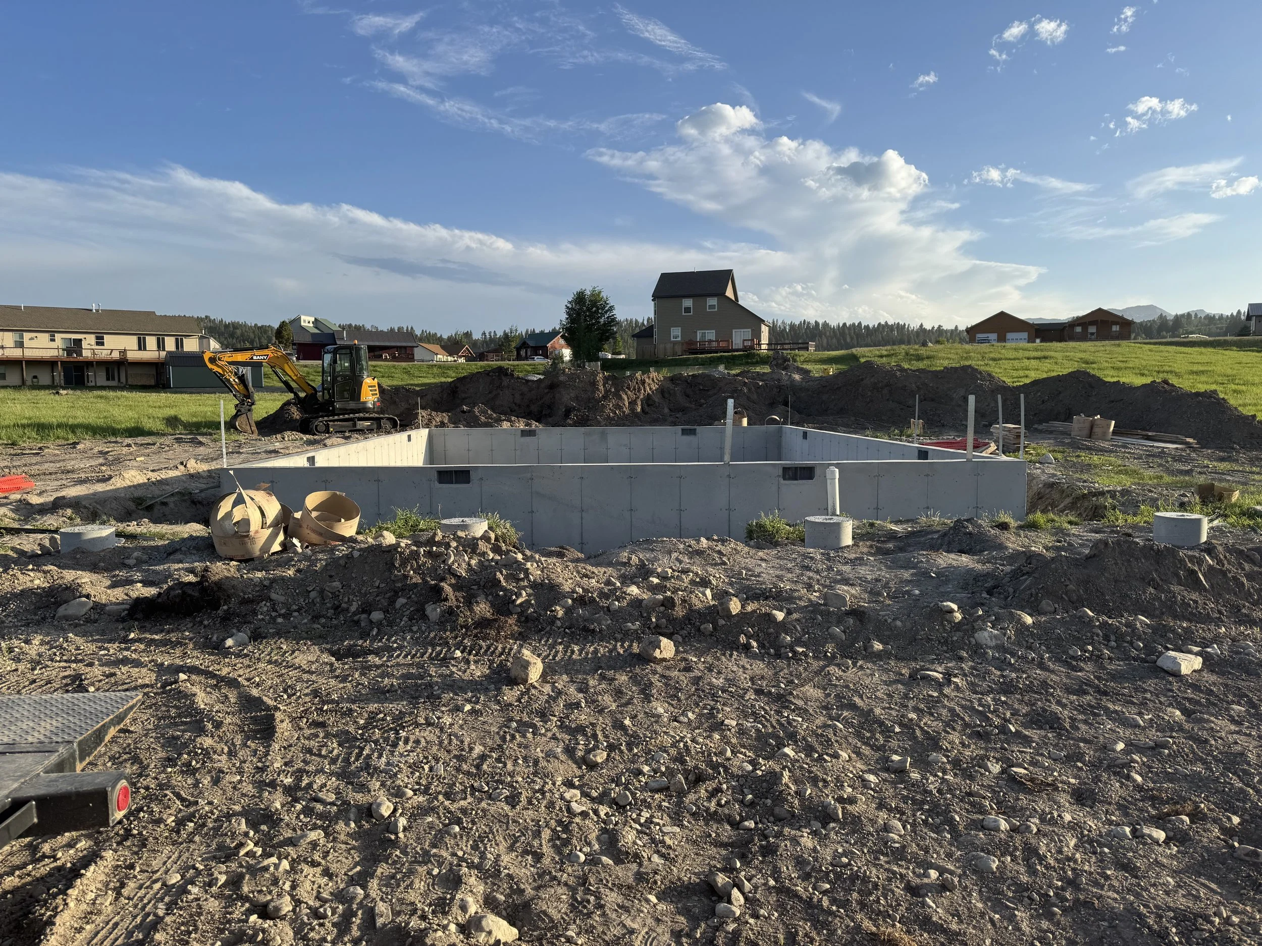 Construction site with a concrete foundation, dirt piles, and a small excavator in a residential area under a partly cloudy sky.
