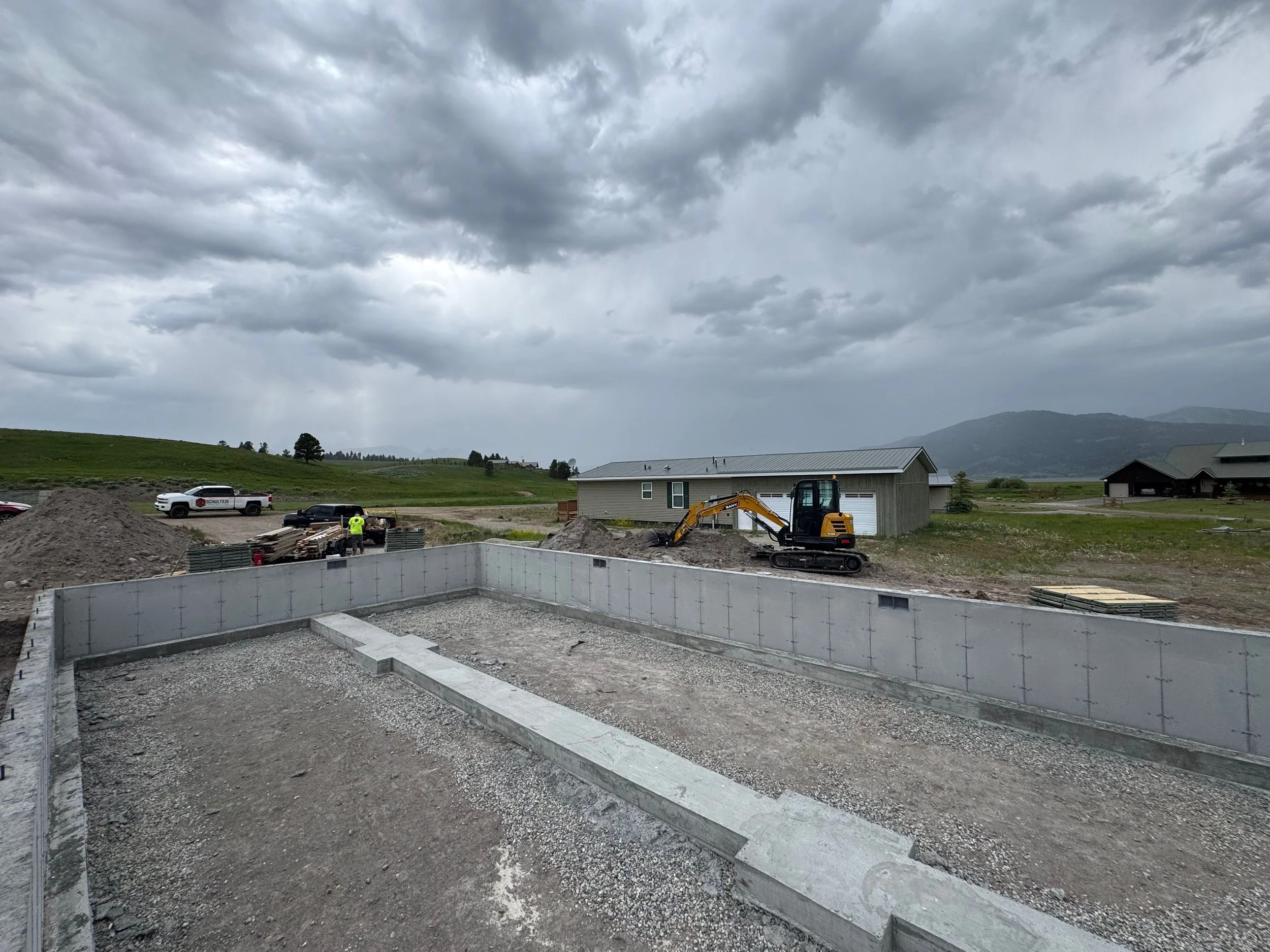 Construction site with a concrete foundation and a small yellow excavator, with construction workers, cars, and a cloudy sky in the background.