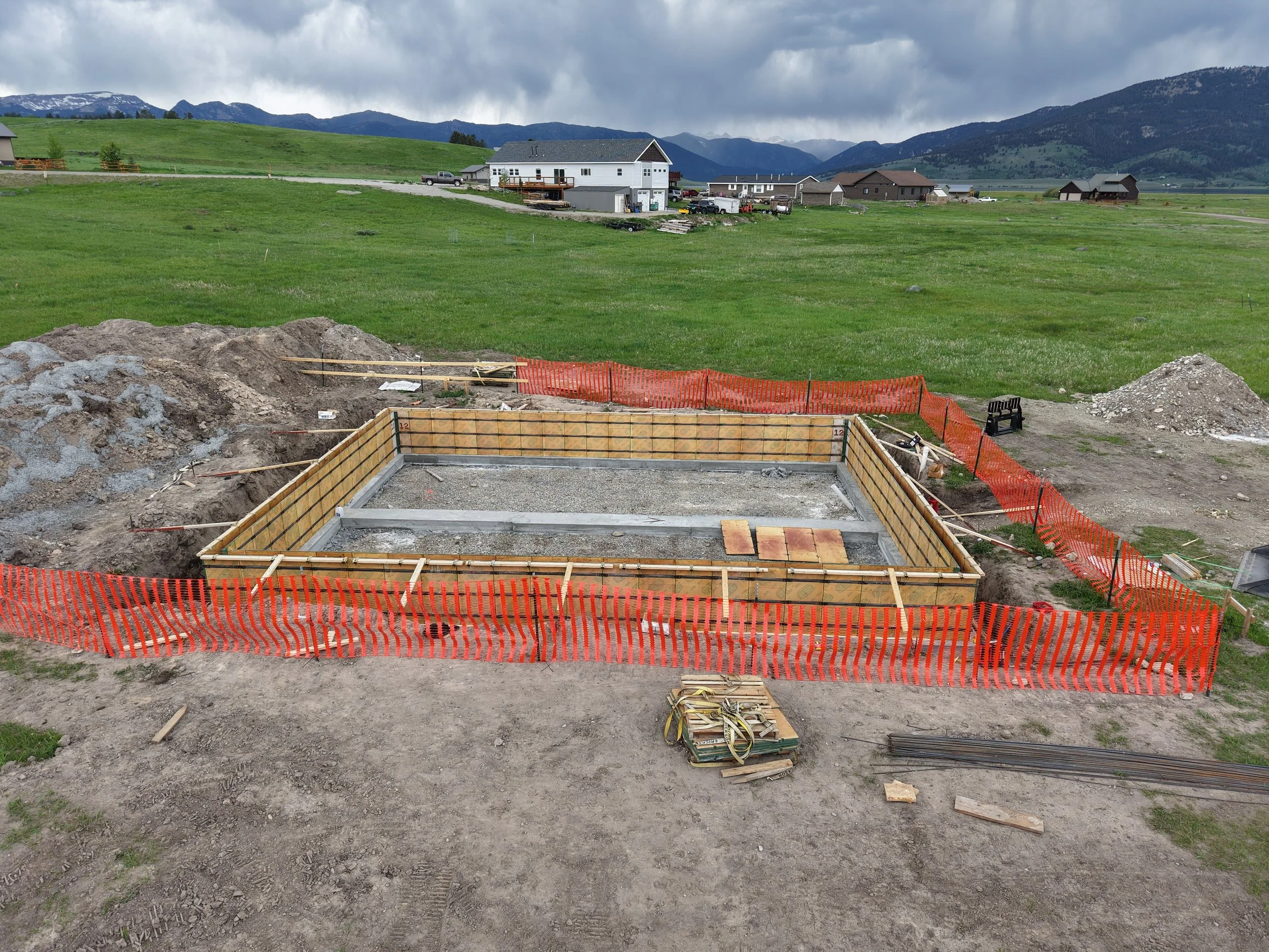 Construction site with a foundation in progress, surrounded by orange safety fencing, in a rural area with green fields and houses in the background.