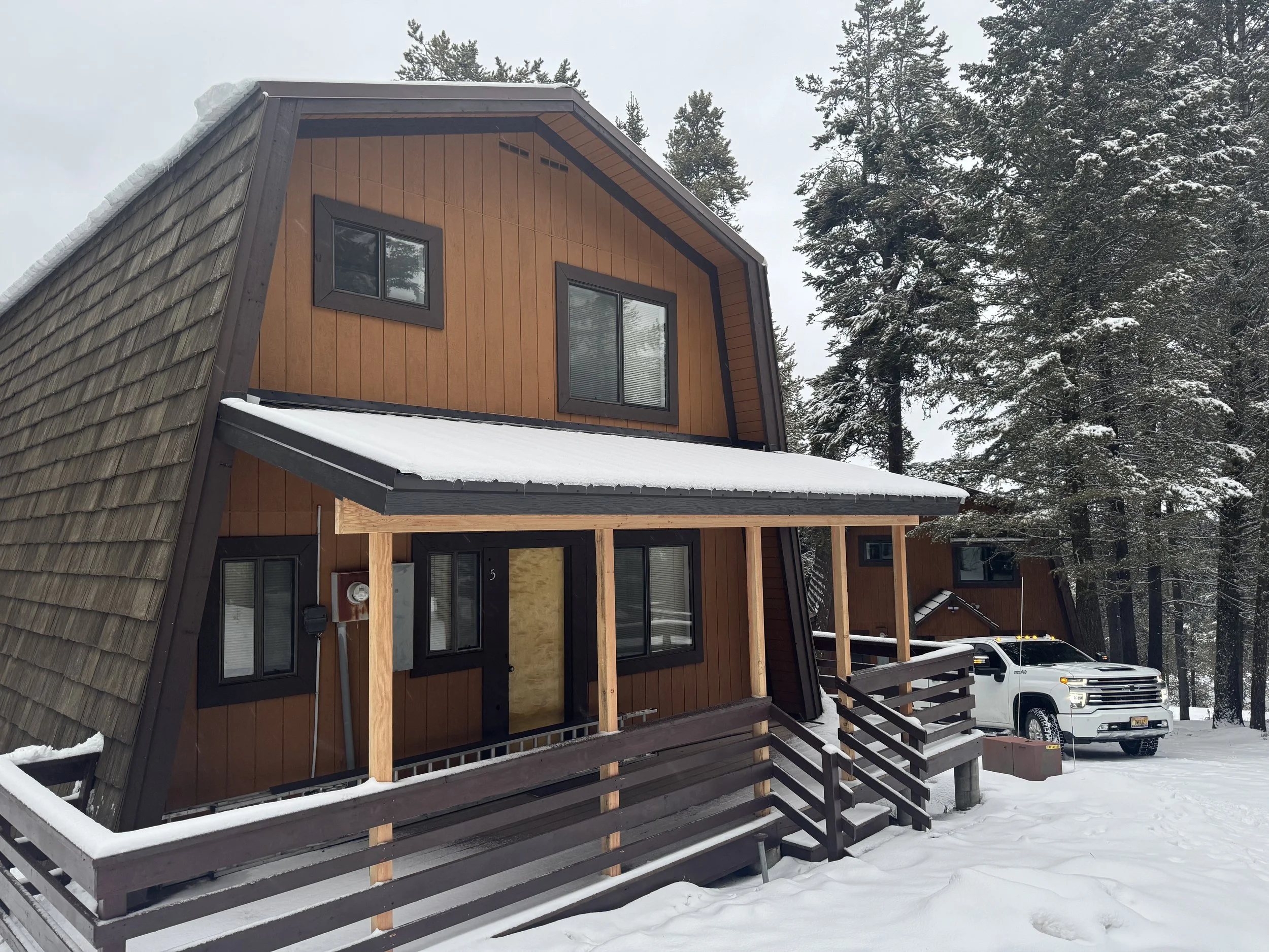 A two-story A-frame house with vertical wood siding and black trim, surrounded by snow and evergreen trees, with a white pickup truck parked outside.