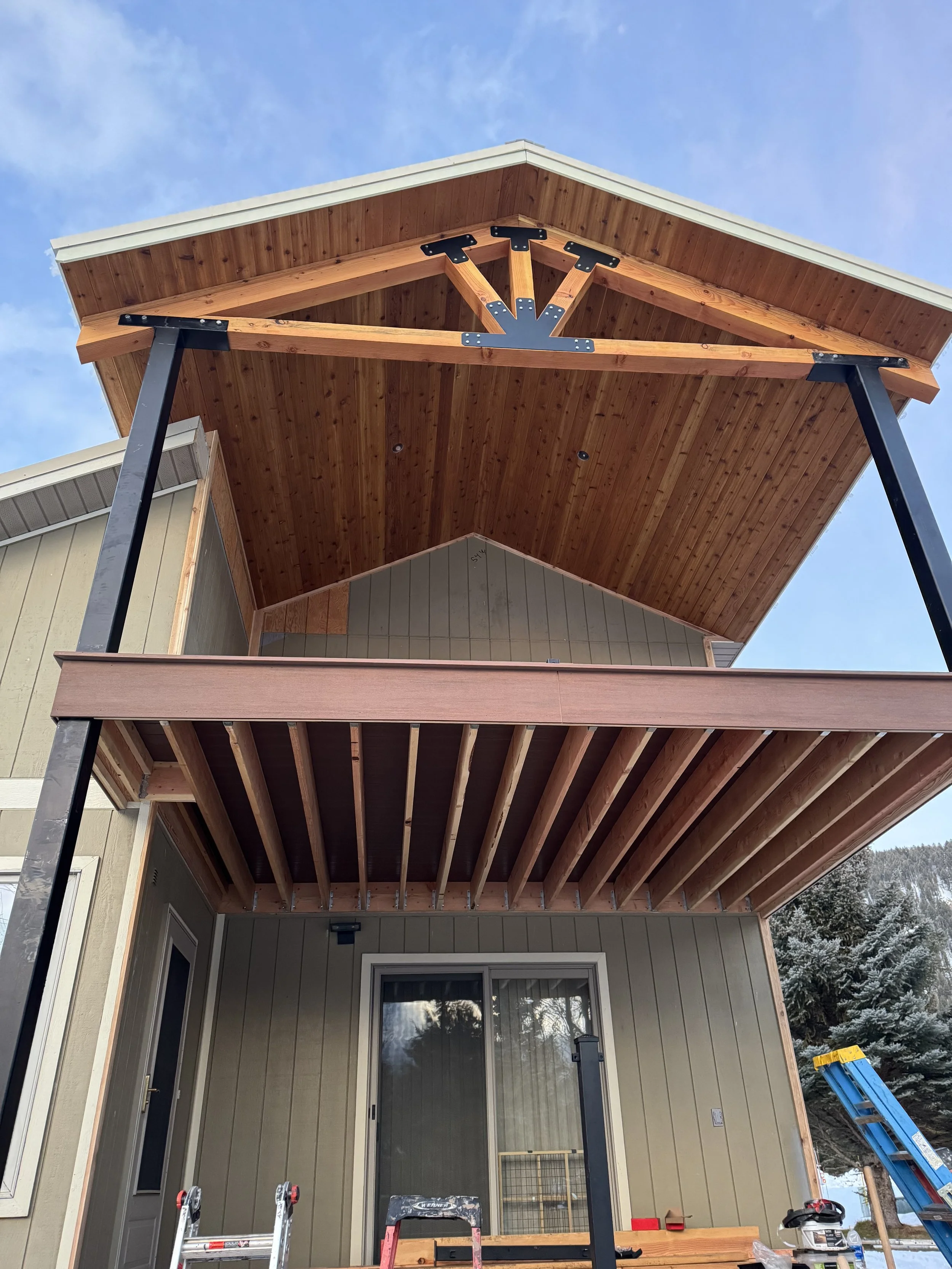 Construction of a two-story house with a covered balcony. The balcony on the second floor has a wooden ceiling and a railing being installed. The house exterior features beige siding and a sliding glass door on the ground floor. There is a ladder and