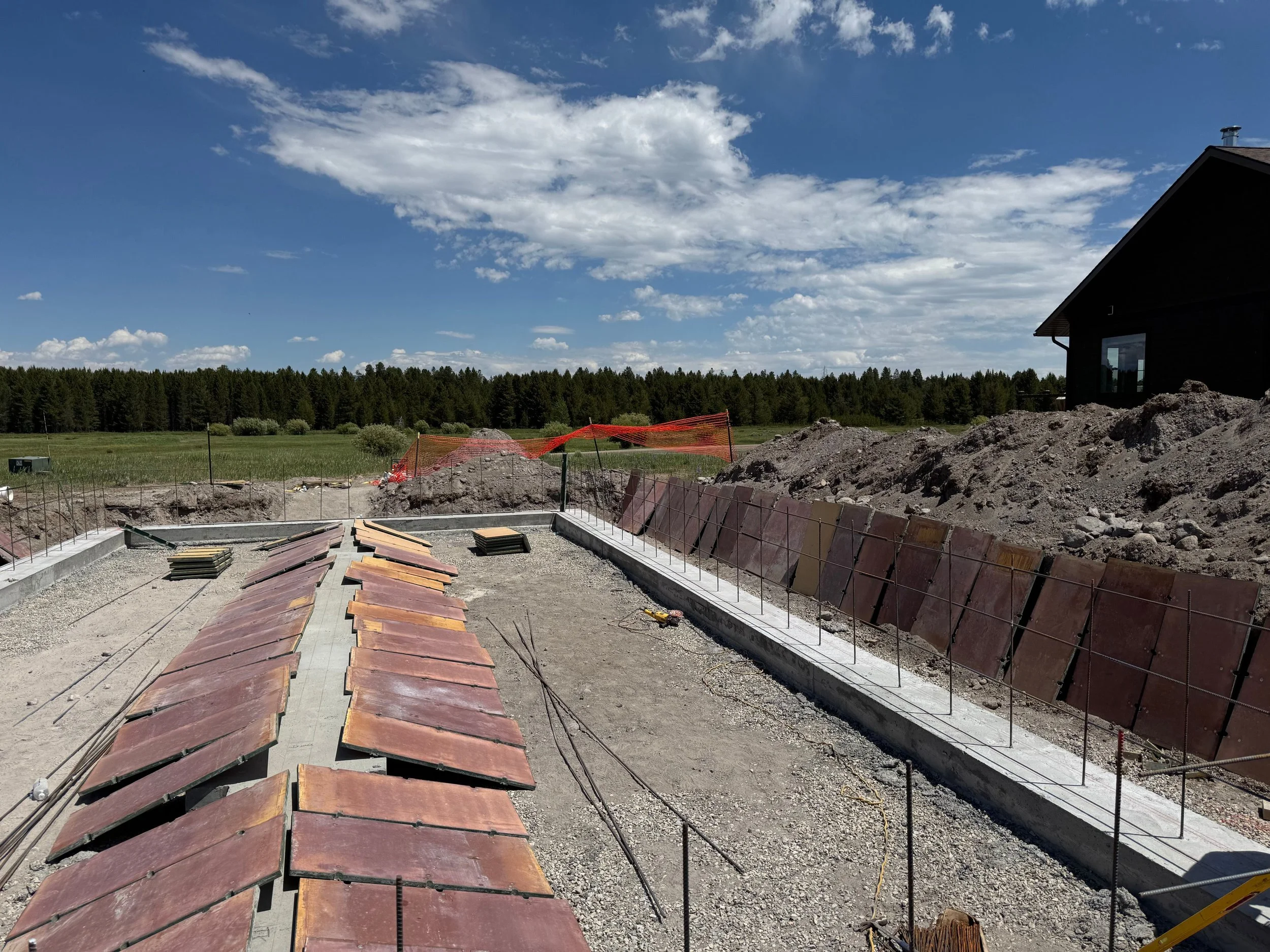 Construction site with concrete foundation, wooden planks, and metal reinforcements, surrounded by dirt mounds and open land, under a blue sky with clouds.