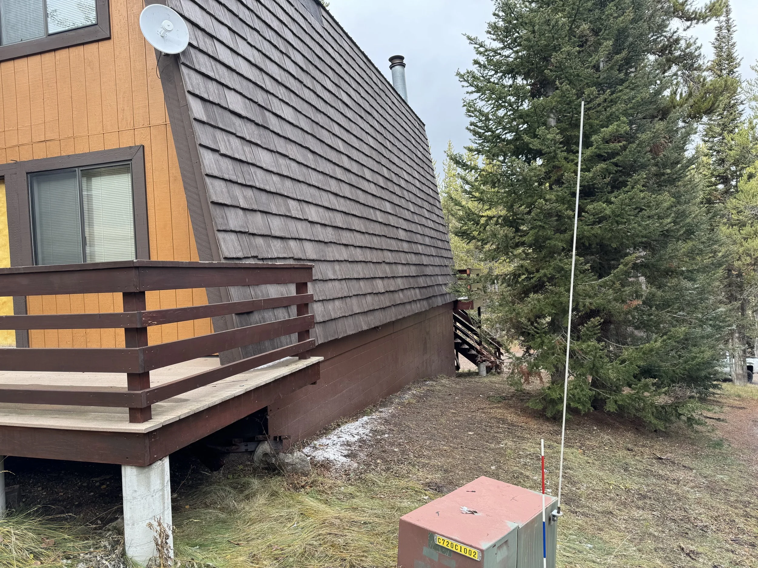 A house with a brown exterior, a sloped roof covered in dark shingles, a small balcony with a brown railing, a window with closed blinds, and a satellite dish. There is a large tree to the right and some electrical equipment on the ground.