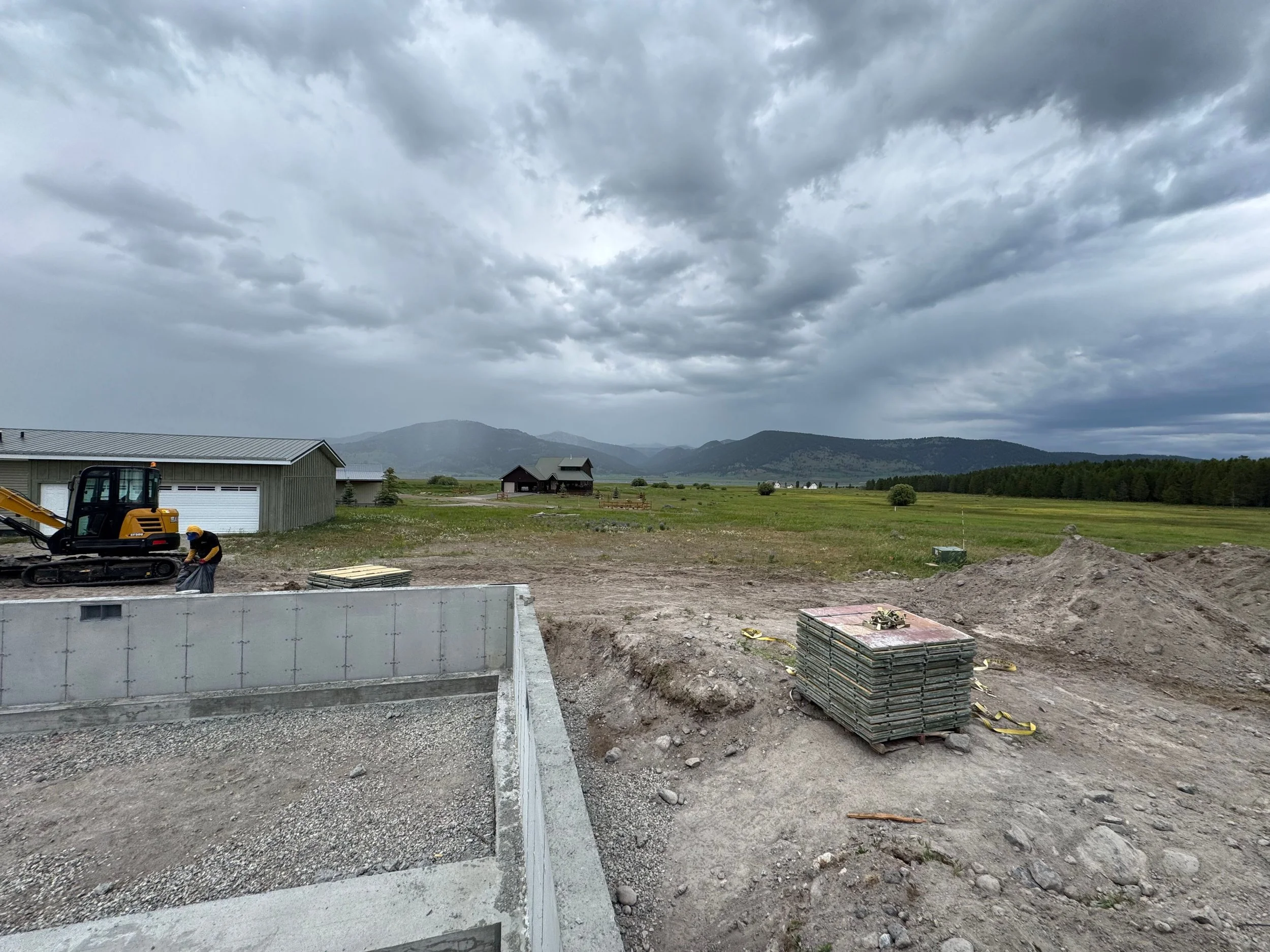 Construction site with excavator, stacks of concrete slabs, and a worker, overlooking a rural landscape with grass fields, houses, and mountains under a cloudy sky.