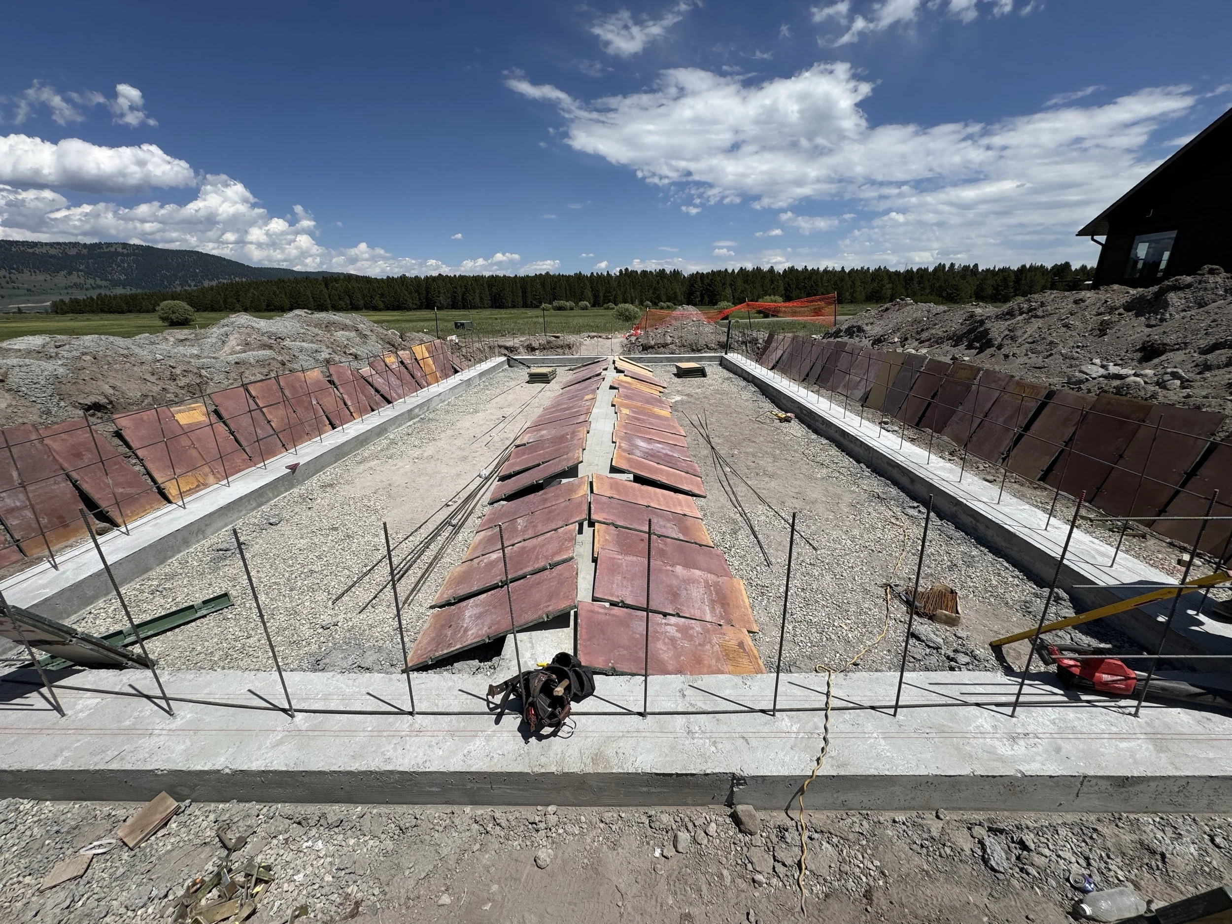 Construction site with a concrete foundation and metal reinforcement bars, with red roofing tiles arranged in the center, surrounded by dirt and gravel, under a partly cloudy sky.