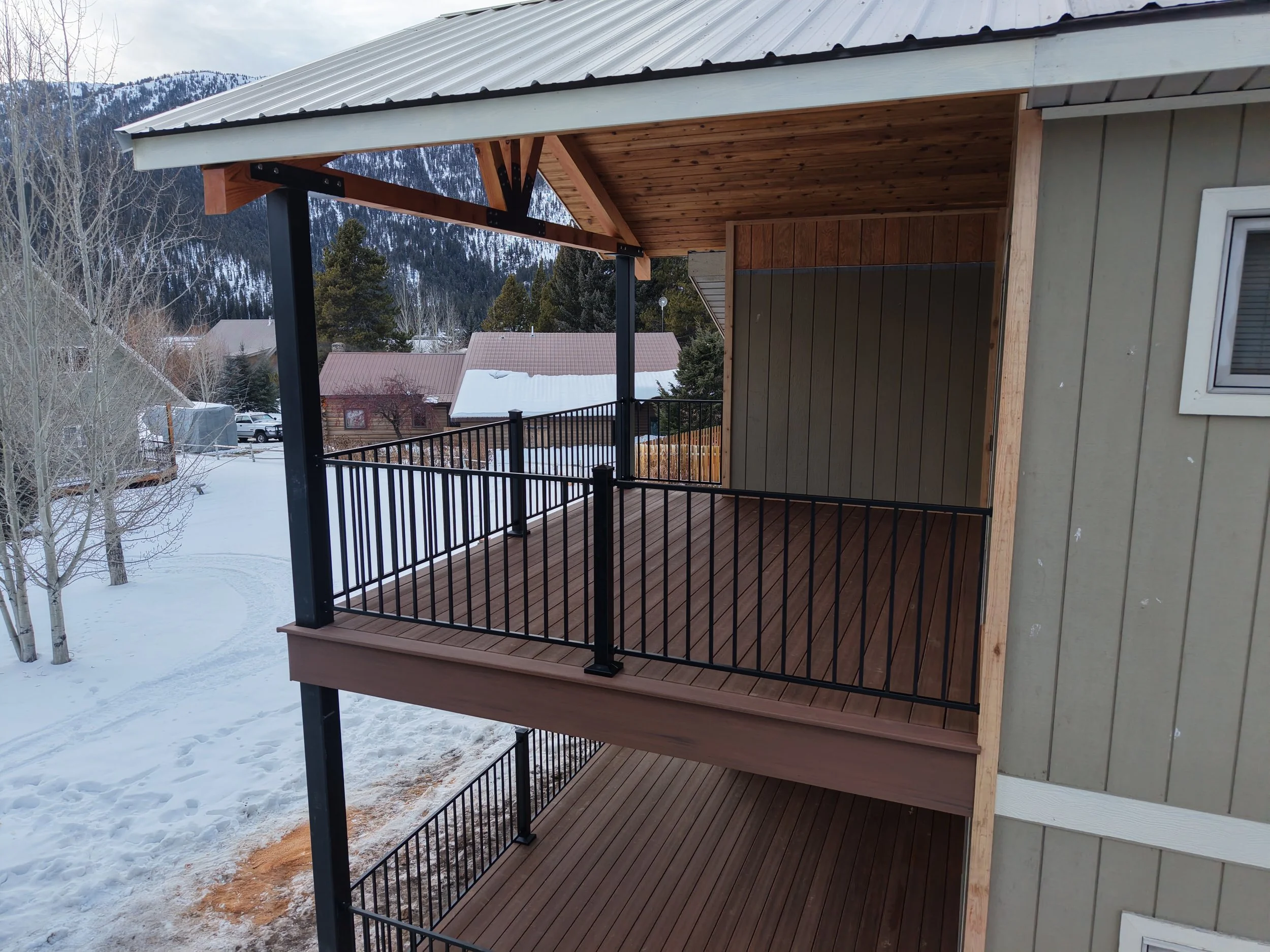 A partially enclosed outdoor balcony with black metal railings and wooden flooring, attached to a house with green exterior siding and a small window, with snow-covered landscape and mountains in the background.