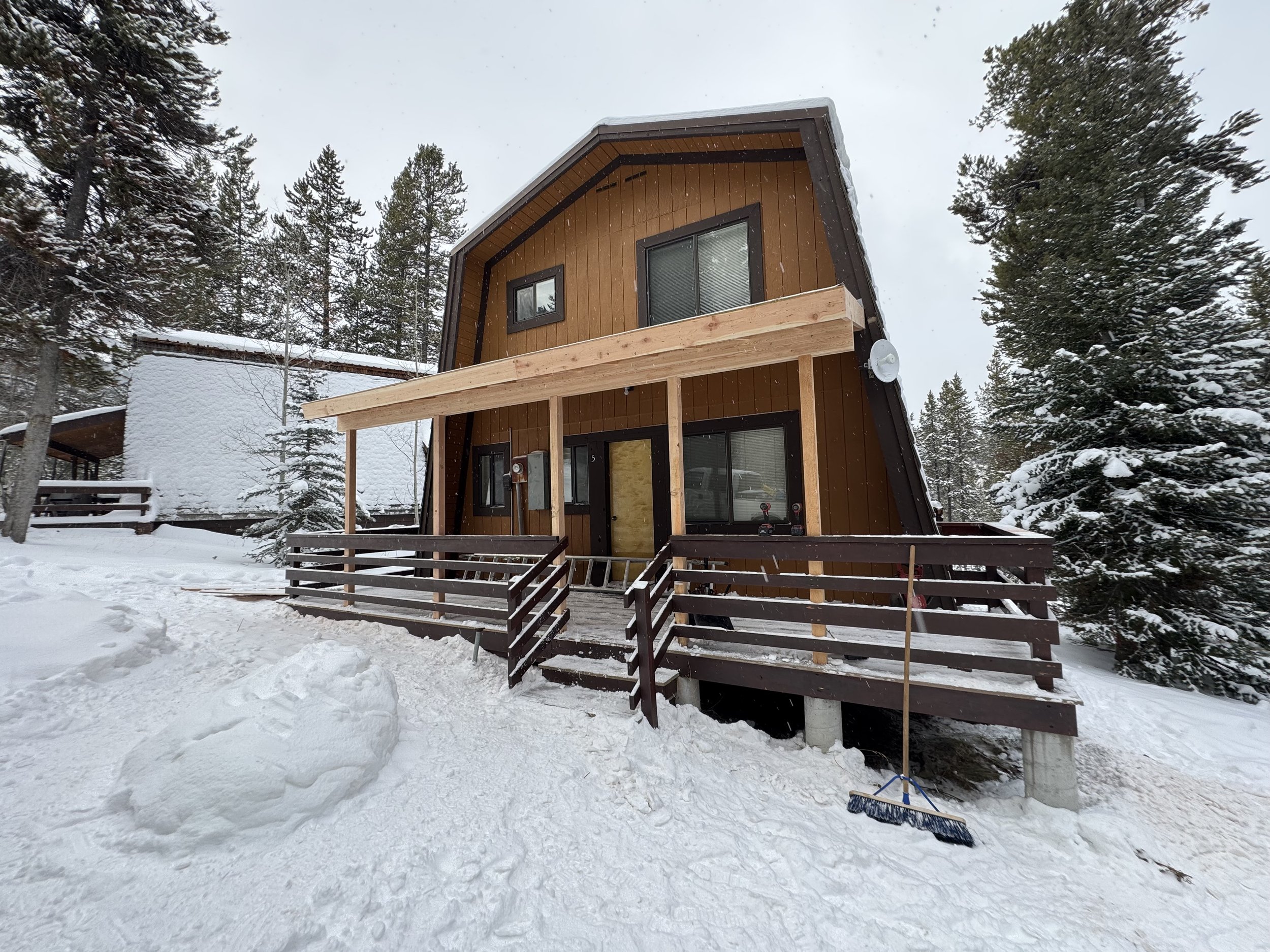 A wooden A-frame house with a front porch and a snow-covered yard, surrounded by tall pine trees on a snowy winter day.