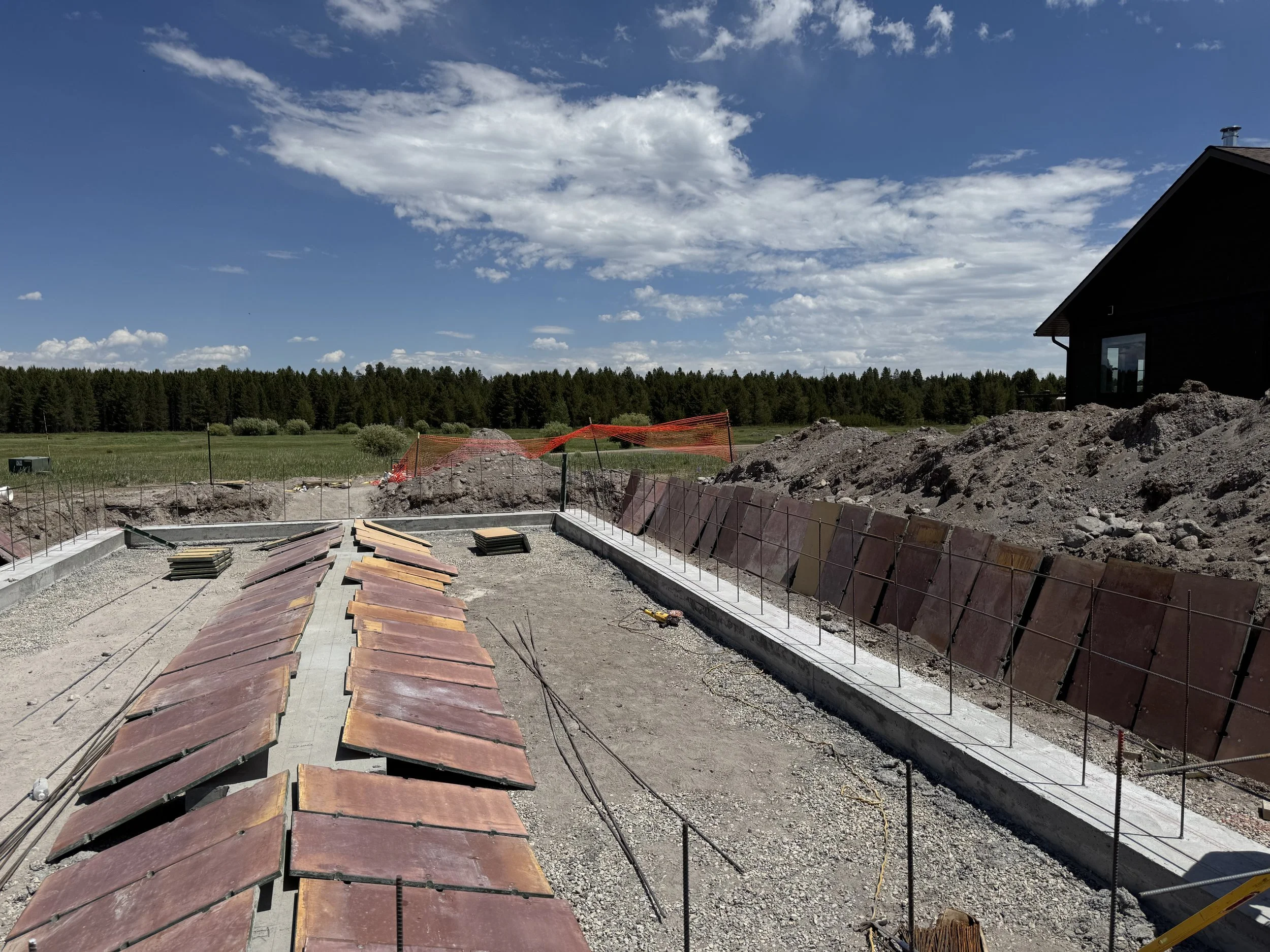 Construction site for a building foundation with trenches, rebar, and tiles, under a partly cloudy sky with a rural landscape and a house in the background.