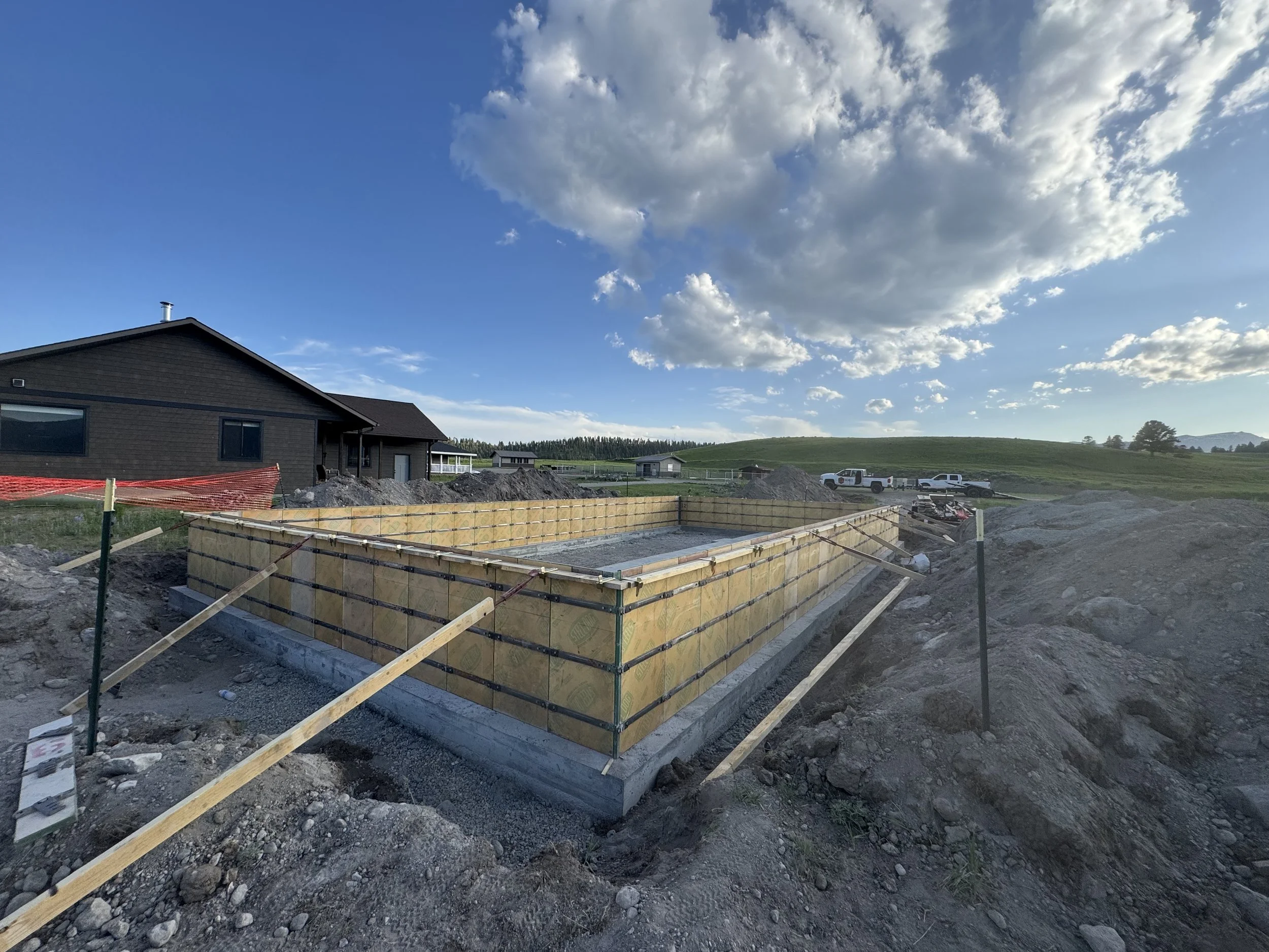 Construction site with a foundation and framing for a new building in a rural area under a partly cloudy sky.
