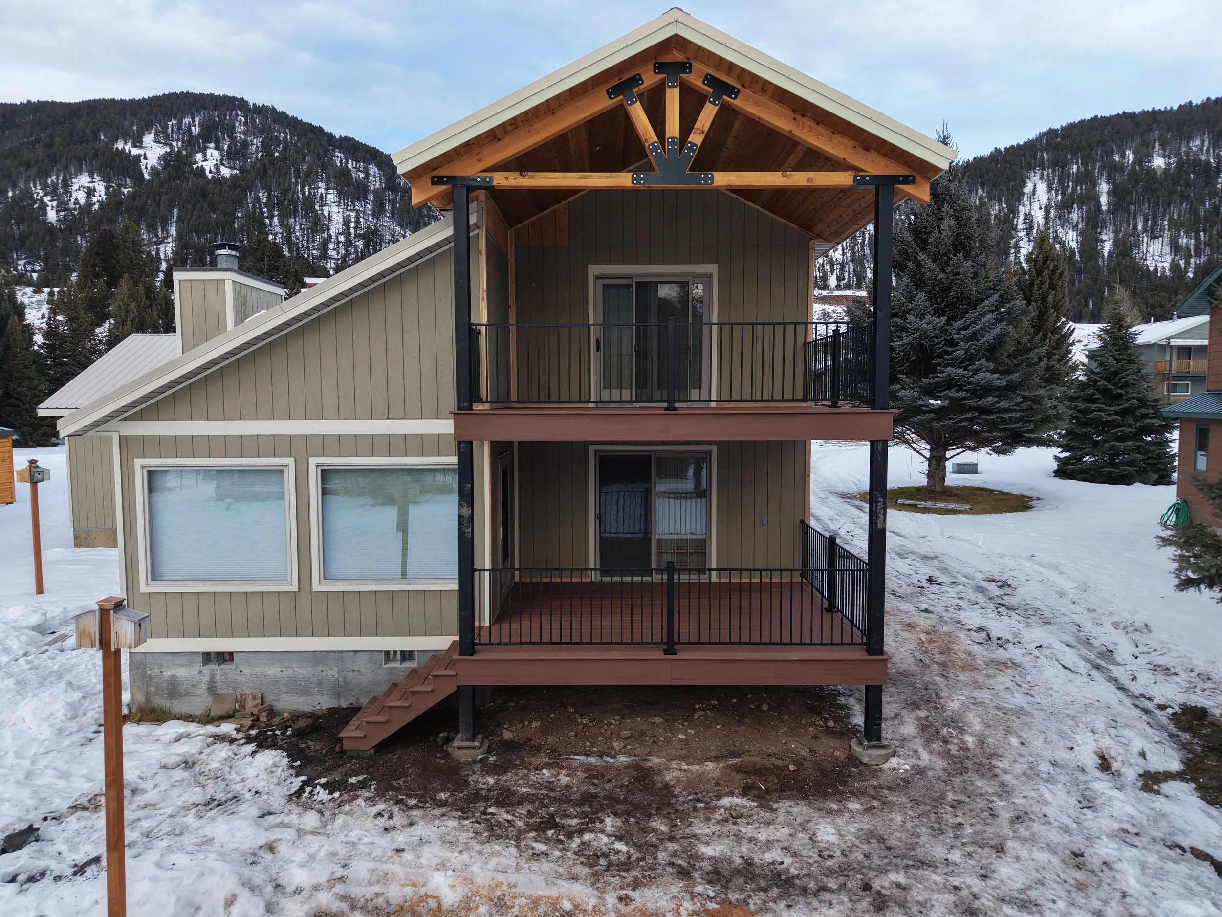 A two-story house with beige siding, a sloped roof, and two balcony decks with black metal railings, set against a snowy mountain landscape.