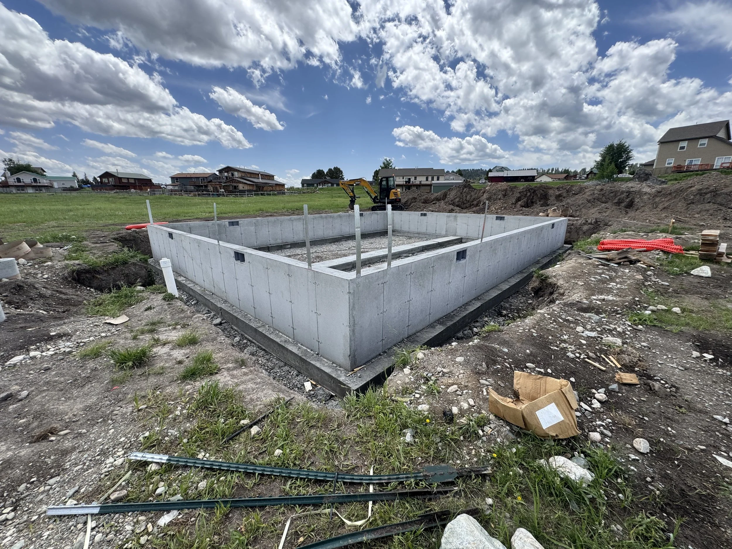 Construction site with a concrete foundation for a building, a small excavator, and a partly cloudy sky.
