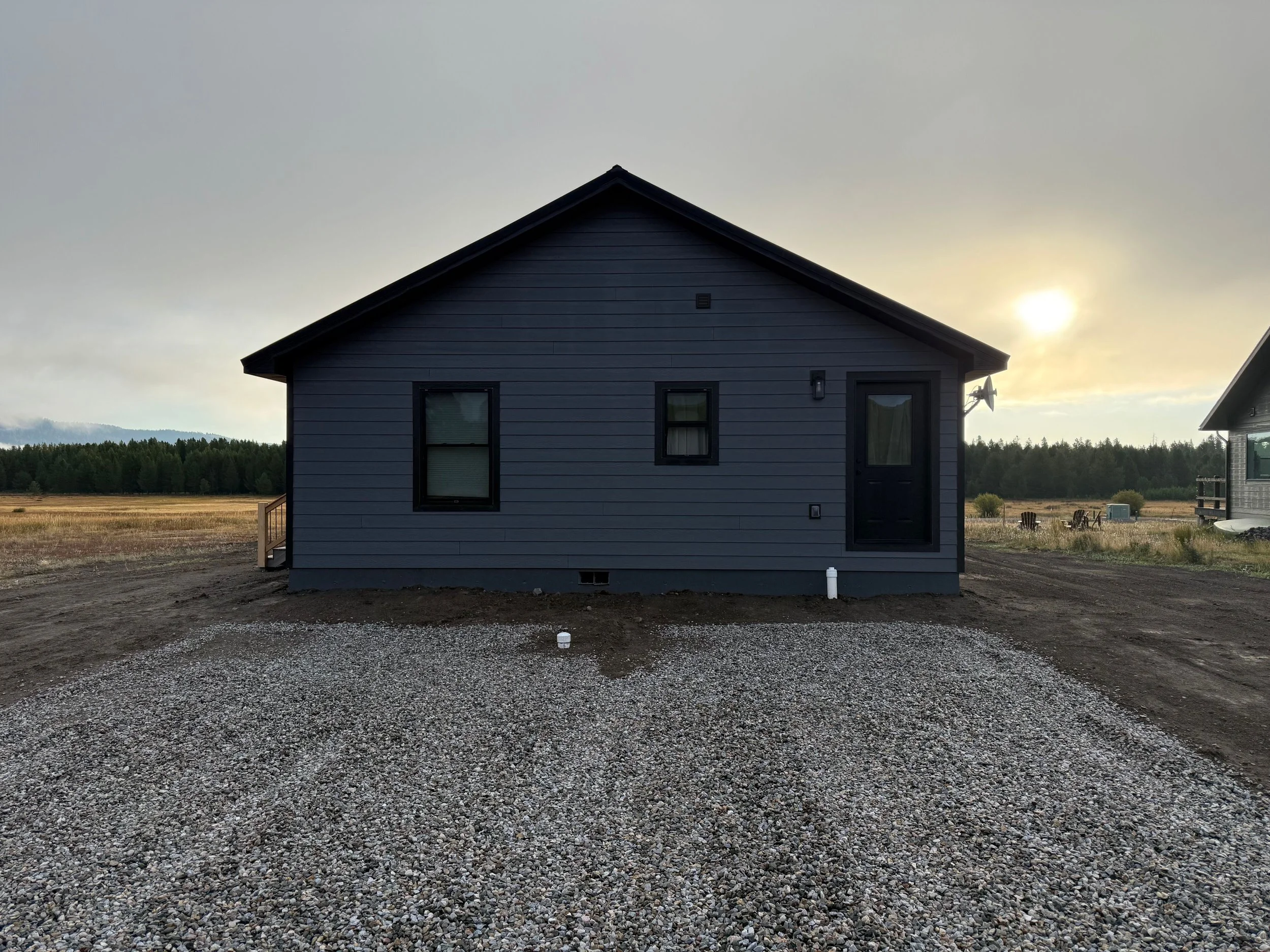A dark blue house with black window frames and a black front door, set in a rural area with a gravel driveway and open fields, under a cloudy sky with the sun partially visible near the horizon.