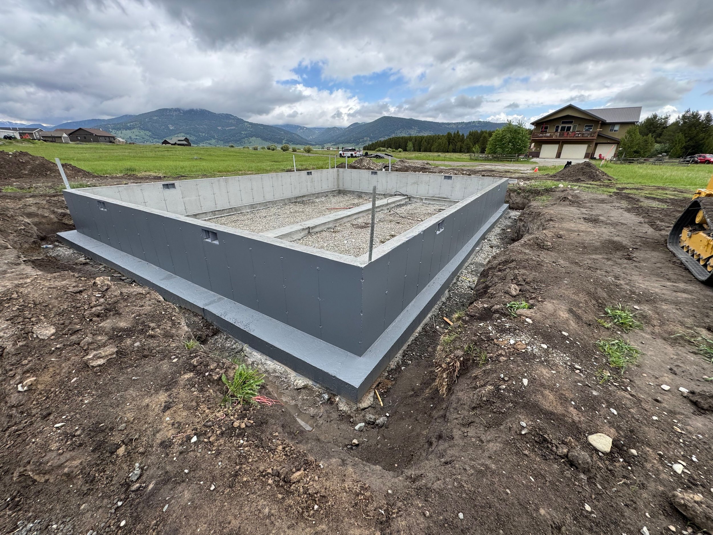 Construction site with empty foundation, gravel base, surrounding dirt, and mountains in the background under cloudy skies.