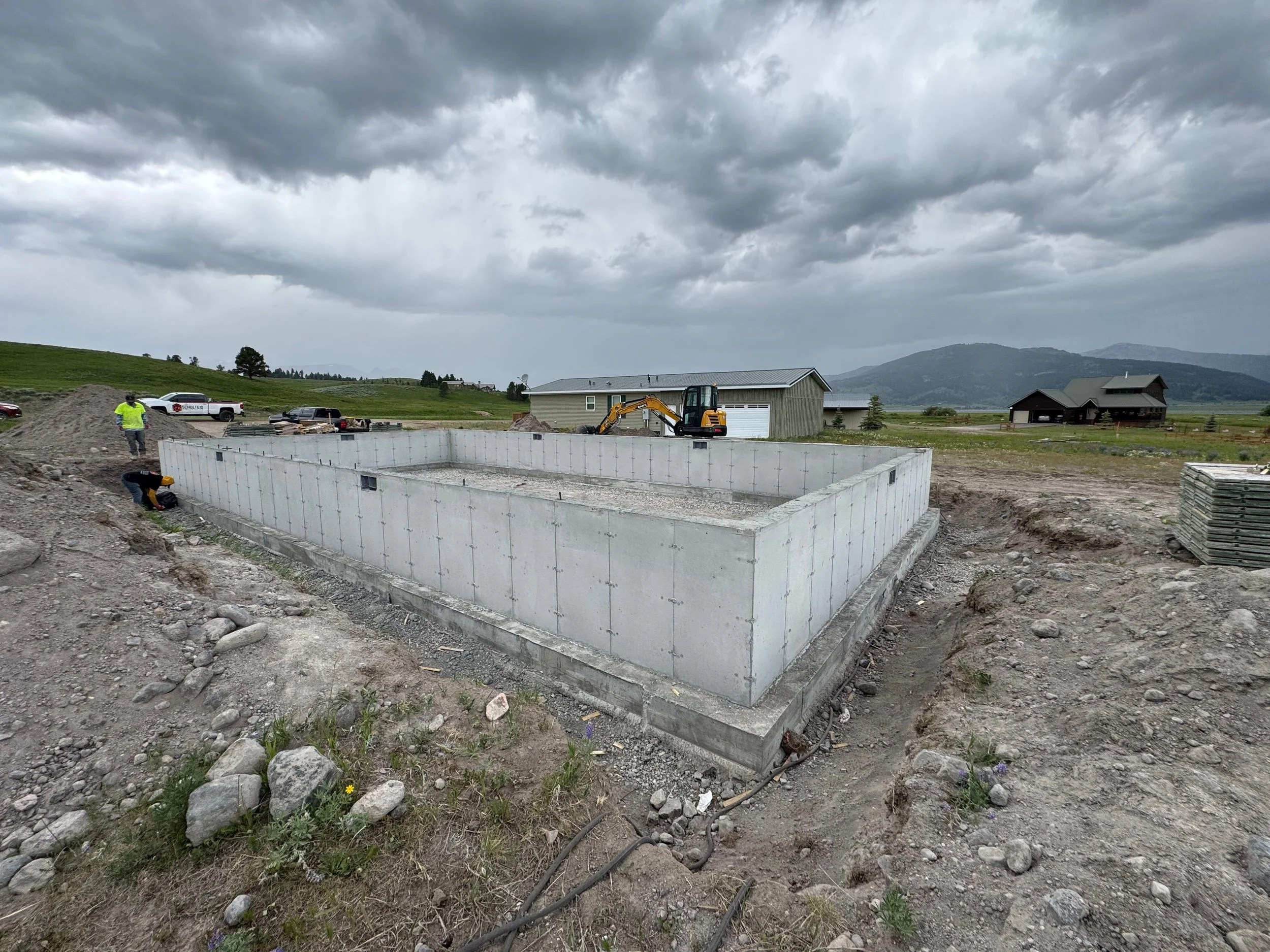 Construction site with concrete foundation, two workers, a small excavator, and a few parked cars in the background, under cloudy sky.