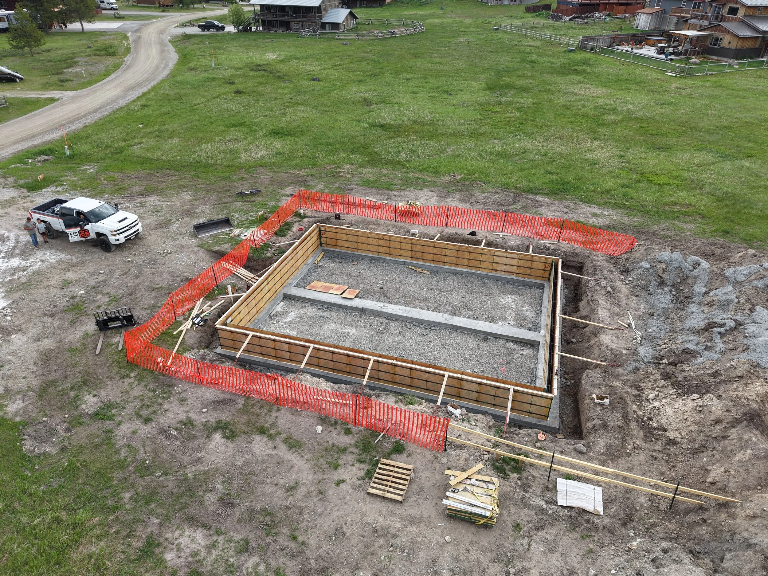 Construction site with a foundation being built, surrounded by orange safety fencing, on a grassy terrain with a dirt road and nearby houses visible in the background.