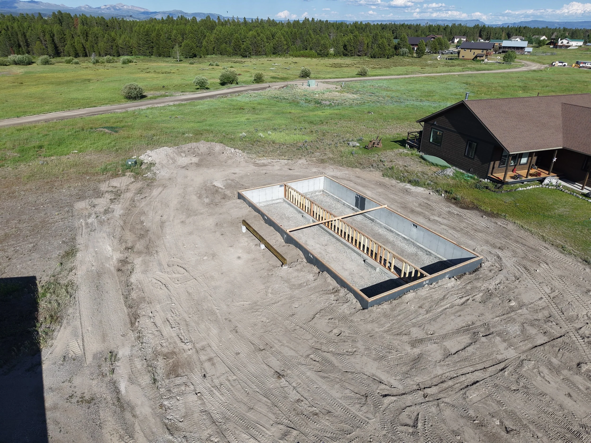 Construction site with a concrete foundation, wooden framing, and dirt surrounding area, with a house and road in the background.