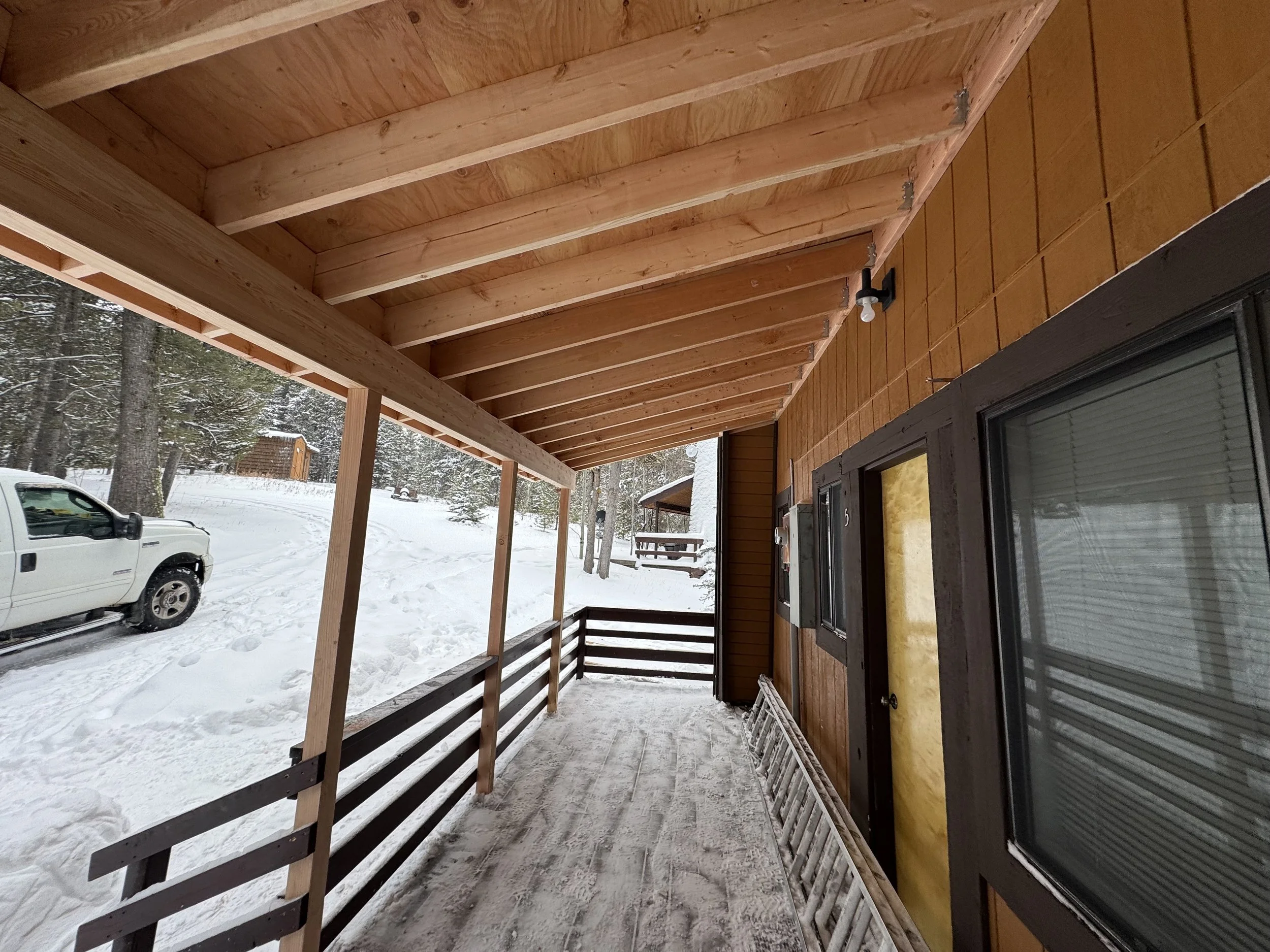 Covered front porch with wooden railings and ceiling, snow-covered ground, parked white pickup truck, and a snowy wooded landscape in the background, part of a brown cabin.