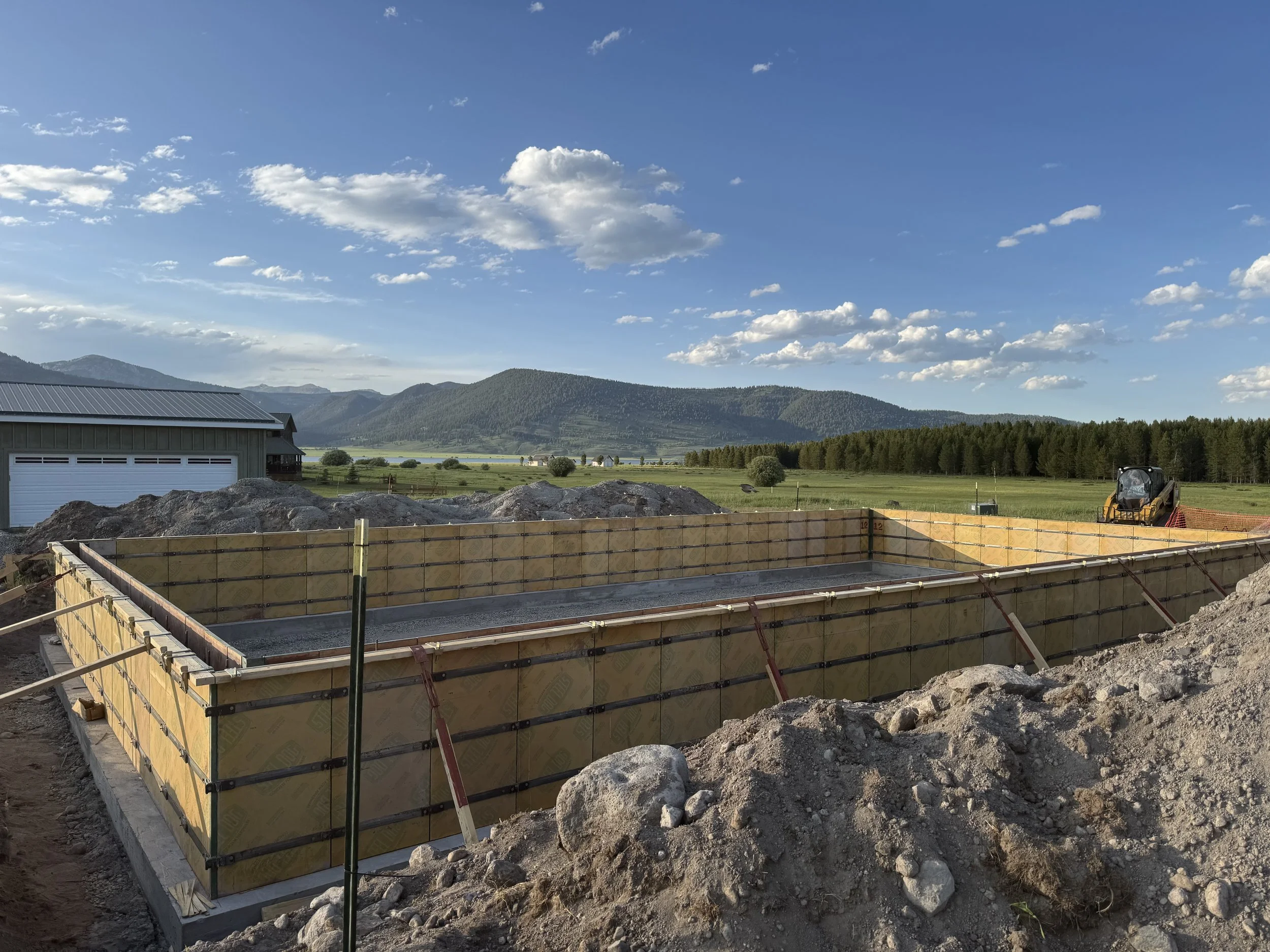 Construction site with a concrete foundation in progress, surrounded by wooden formwork, situated in a rural landscape with green fields, mountains, a small building, and construction equipment under a partly cloudy sky.