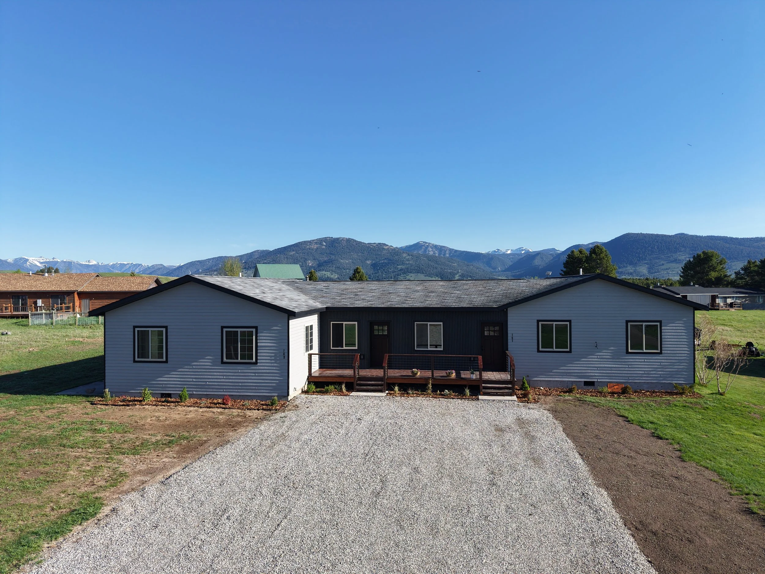 Single-story house with white siding, black trim, and a gravel driveway, surrounded by grassy yard and distant mountains under a clear blue sky.