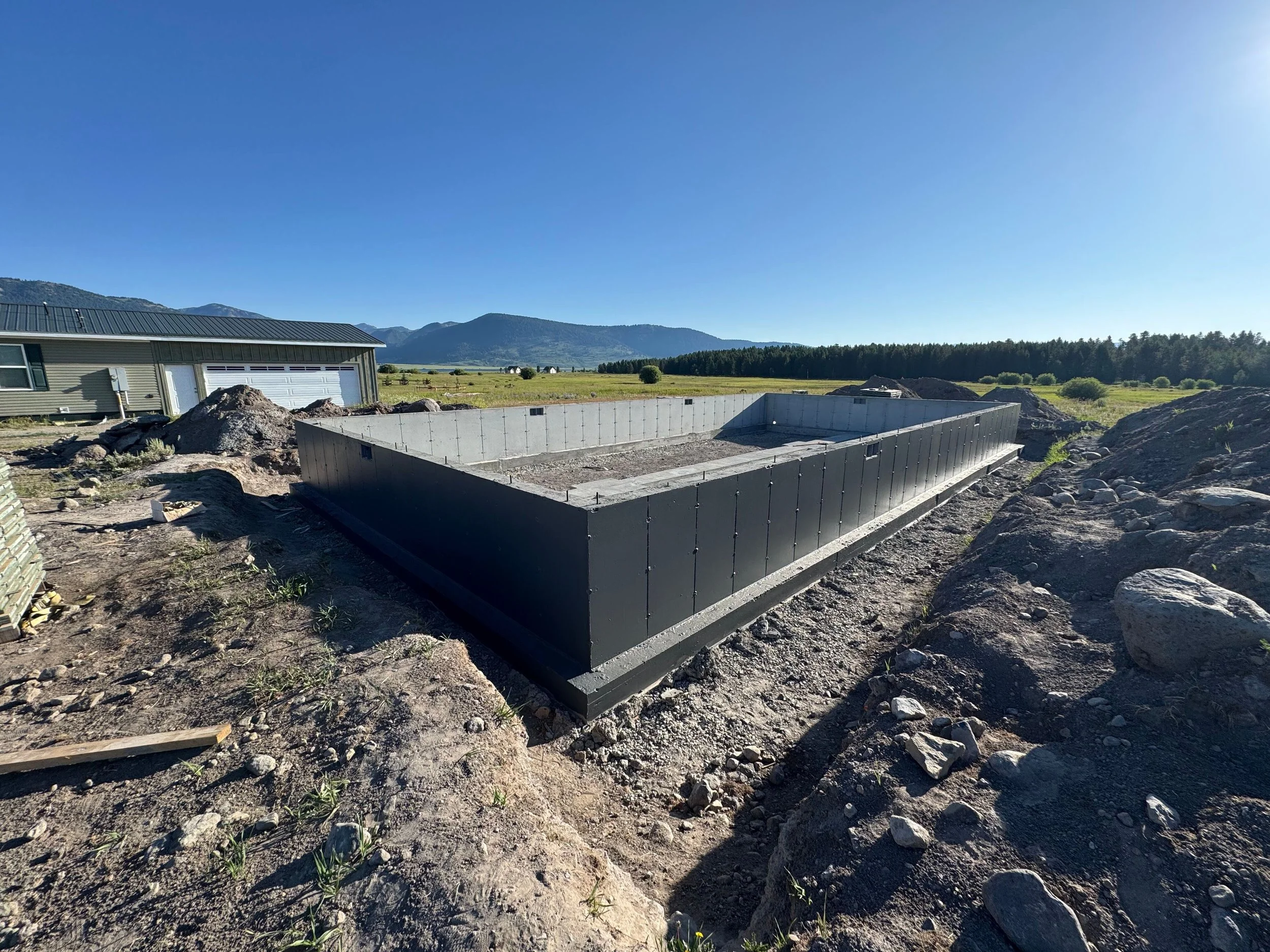 Foundation of a construction site with concrete walls, surrounded by dirt and rocks, with a clear blue sky and mountains in the background.