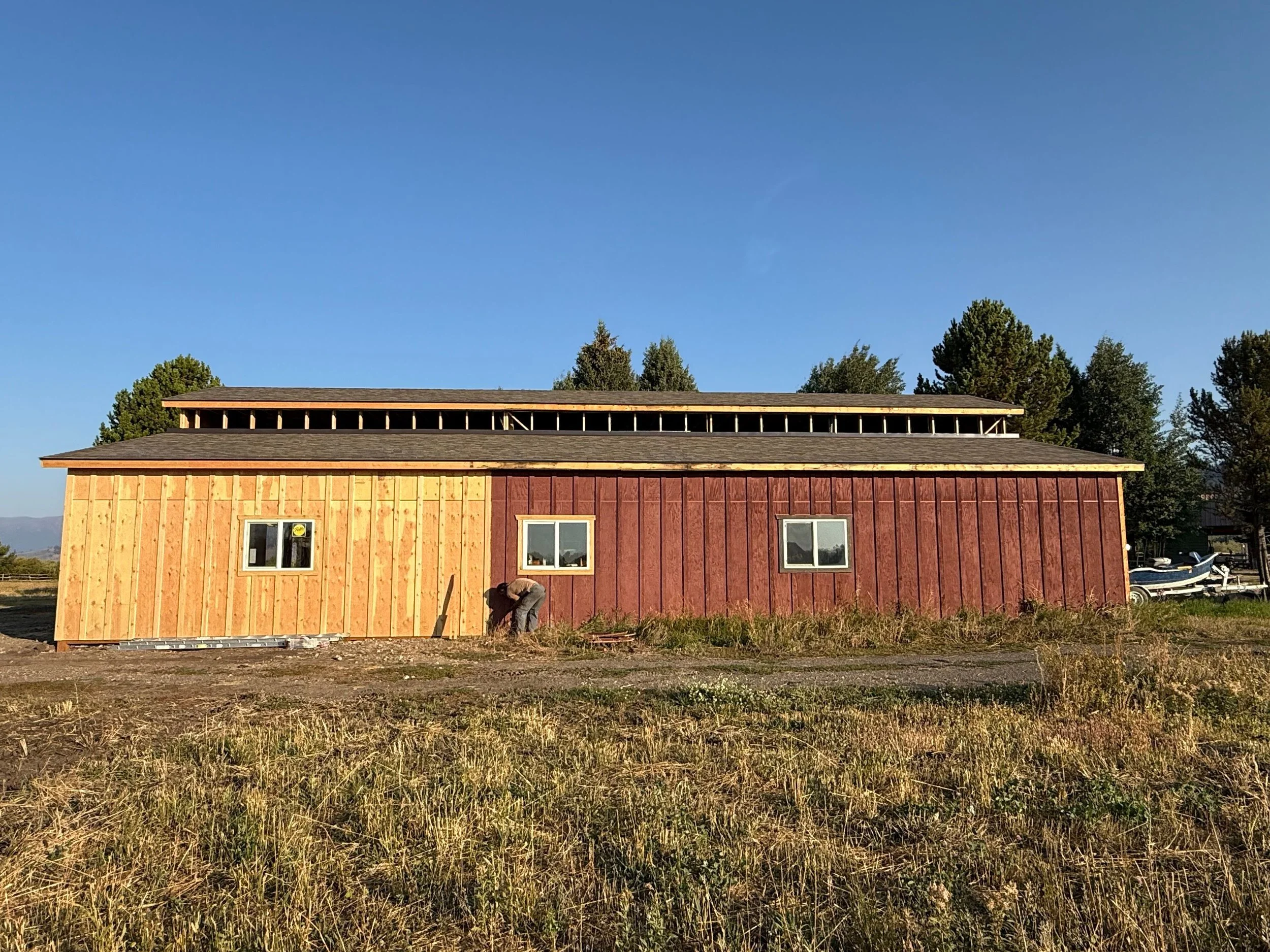 A partially constructed building with wood siding, three windows, and a worker working on the exterior. The building is set on a grassy area, with trees and a boat in the background under a clear blue sky.