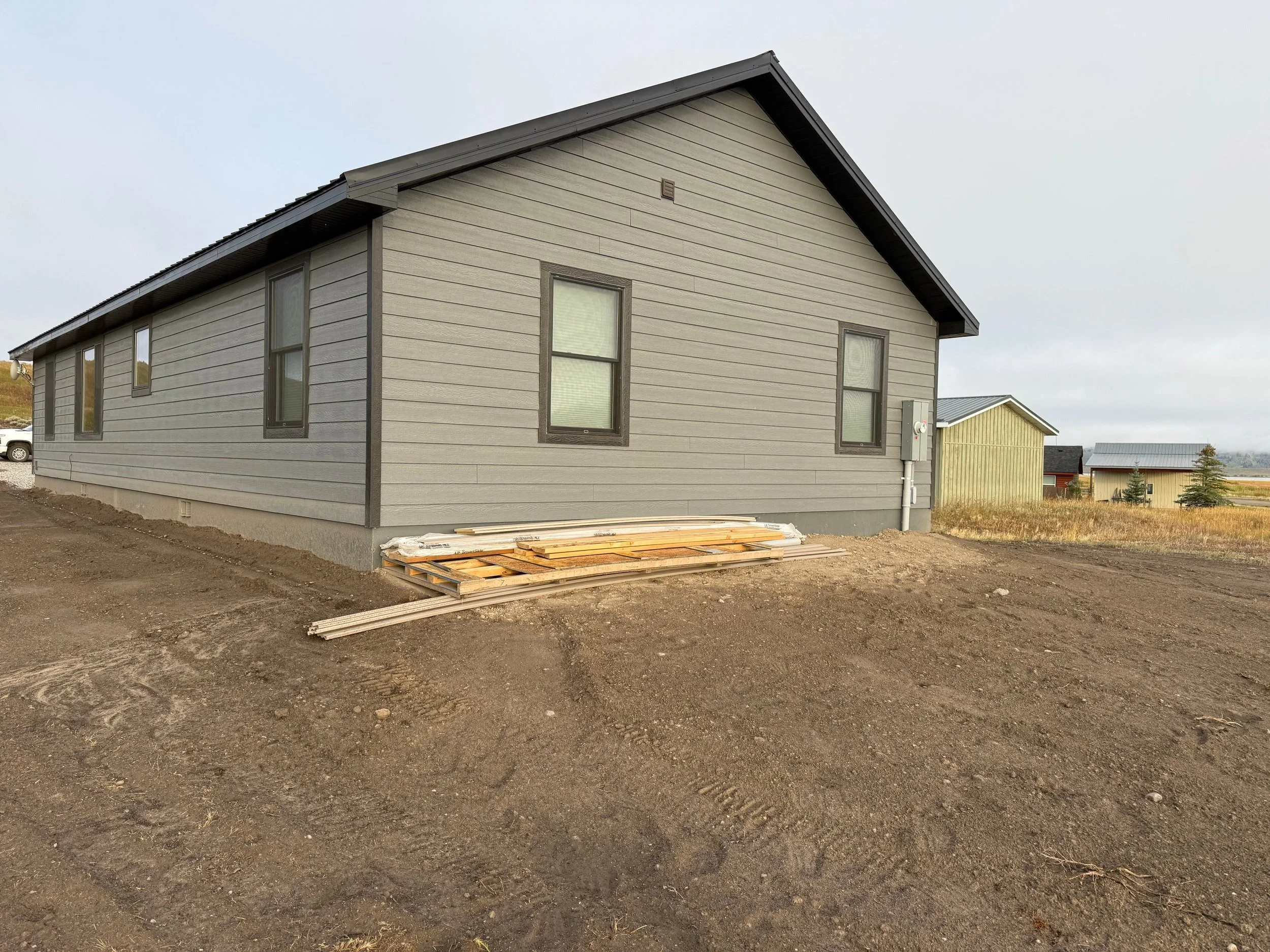 A house under construction with gray siding and three windows on the side facing the viewer, with a small pile of building materials on the ground near the foundation, on a dirt lot with other buildings in the background.