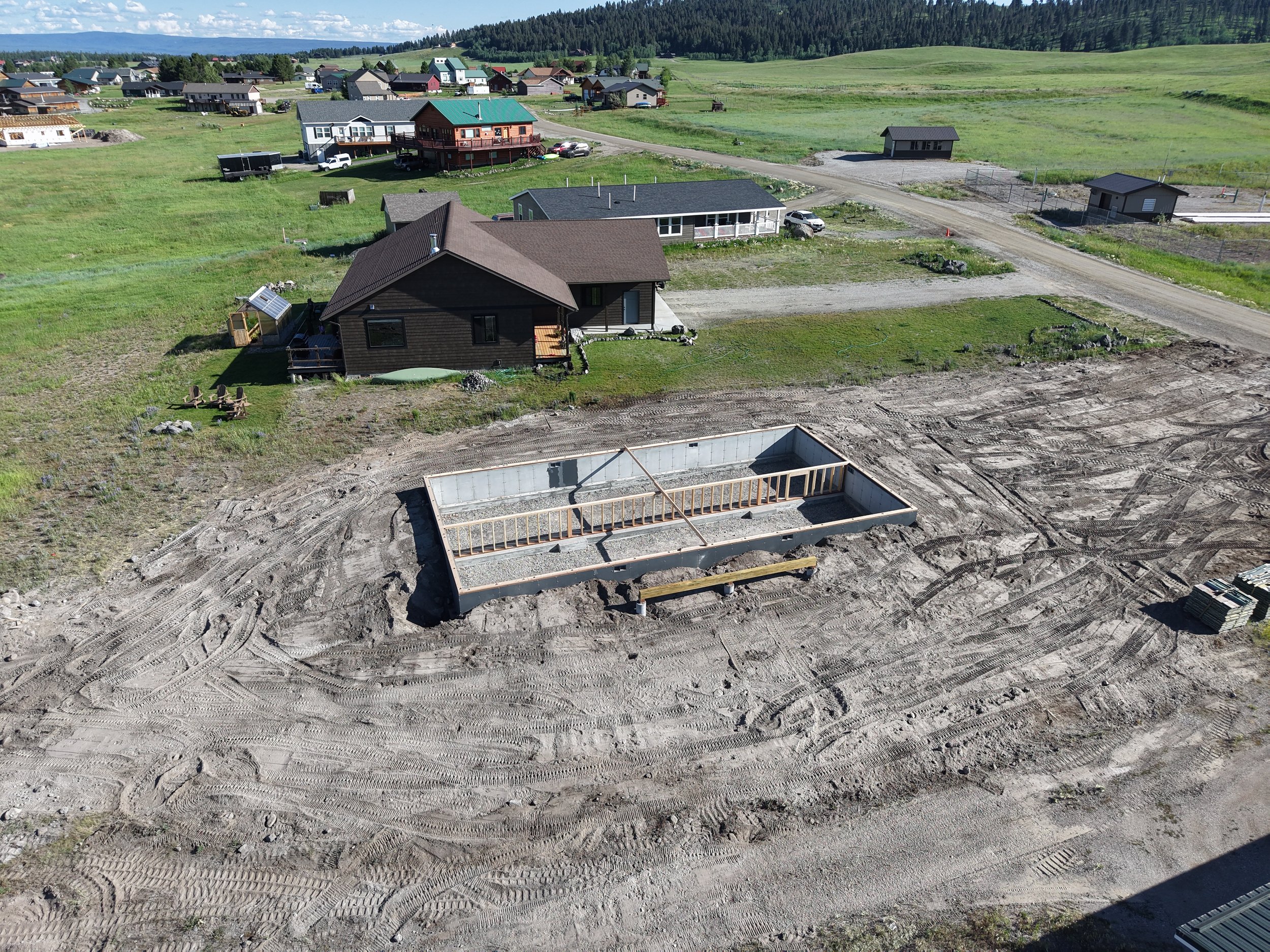 A construction site with a building foundation and wooden framing, surrounded by dirt and tire tracks, in a residential area with houses and green fields in the background.