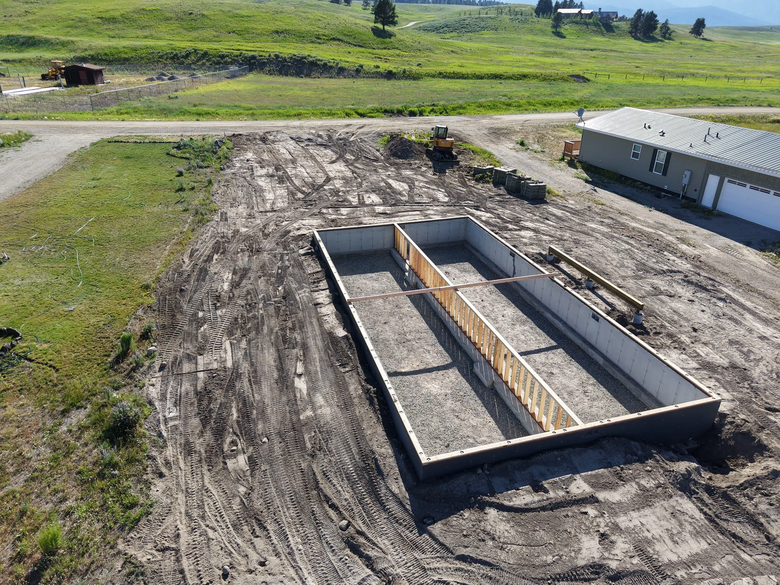 A construction site with a concrete foundation frame, surrounded by dirt and tire tracks, with construction equipment and a house nearby in a rural area with green hills.