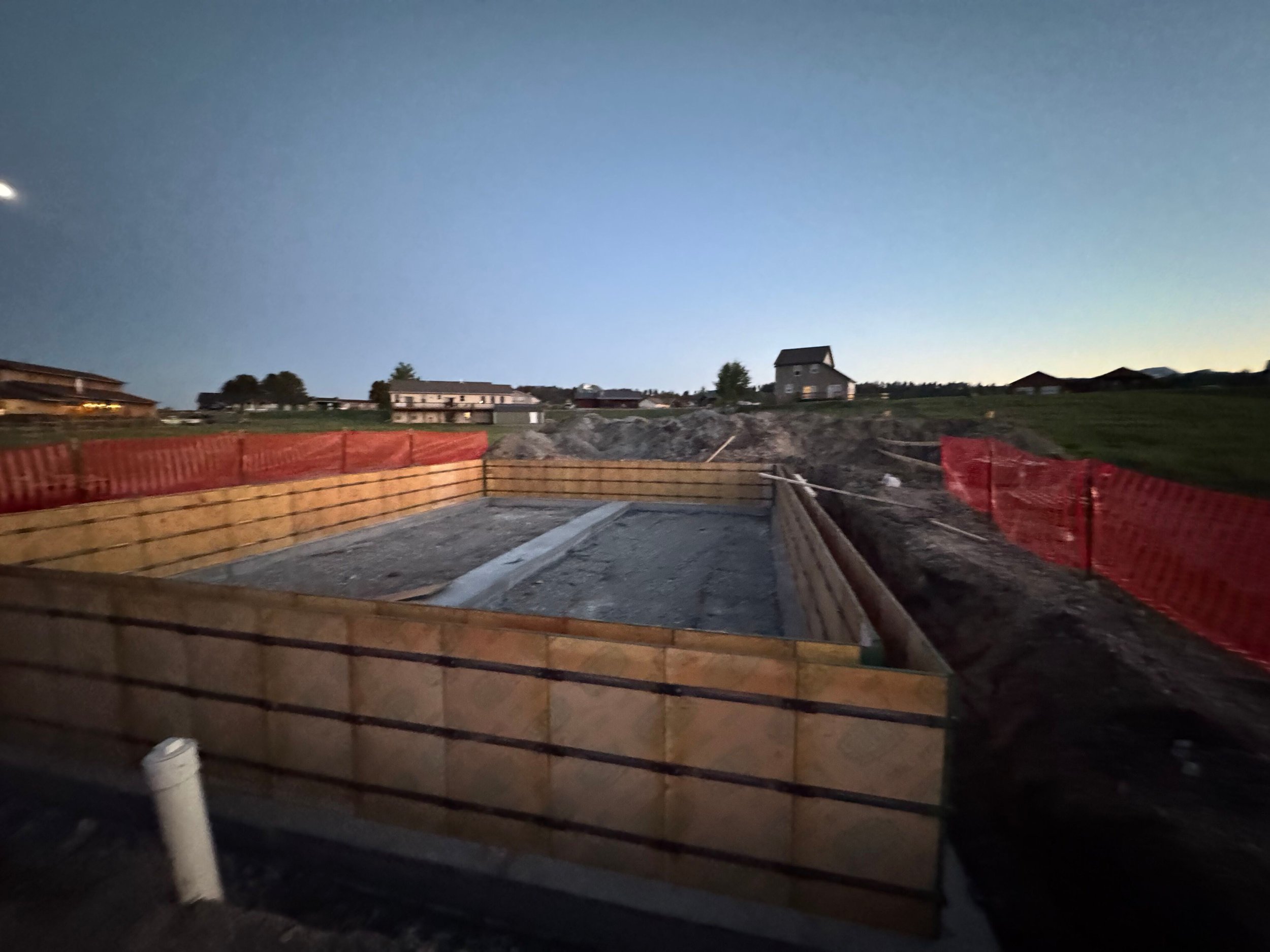 Construction site with wooden framing for a foundation, fenced by red and orange barriers, during early evening or dusk, with houses in the background under a blue sky.