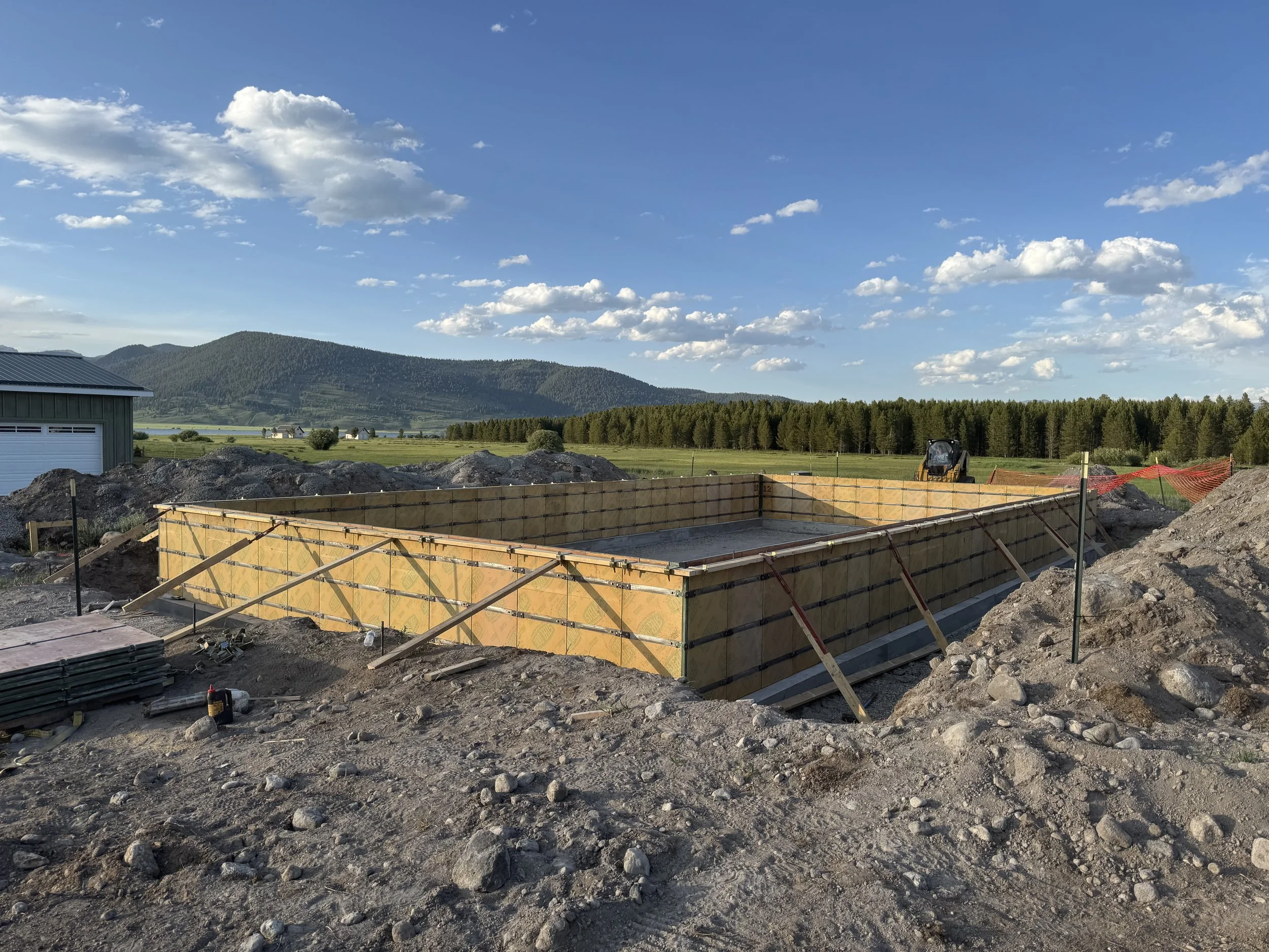 Construction site with a concrete foundation frame, surrounded by dirt and rocks, with mountains, trees, and a blue sky with clouds in the background.