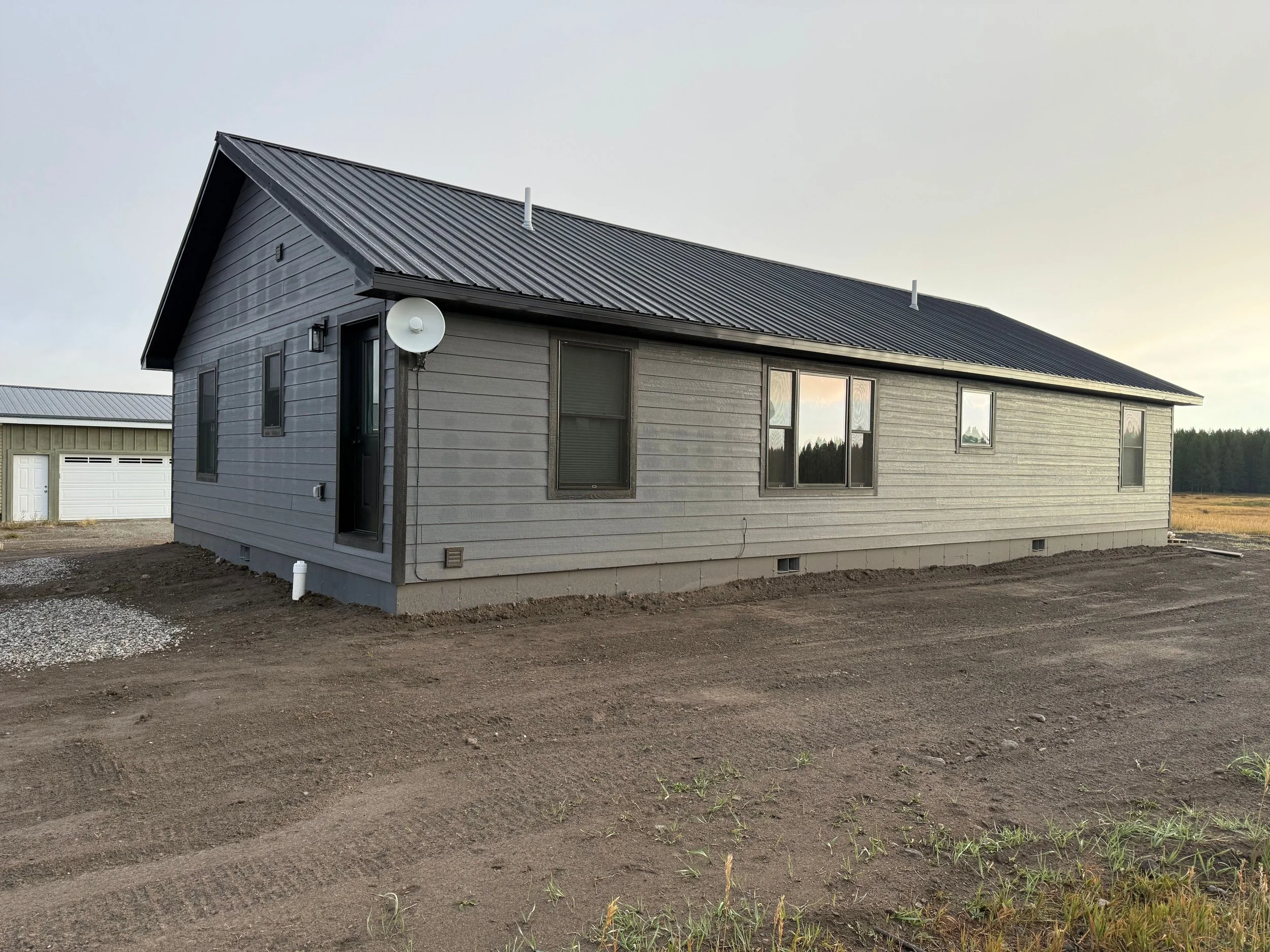 A newly constructed gray house with a metal roof, multiple windows, and a satellite dish, situated in a rural area with some dirt and minimal grass in the foreground.