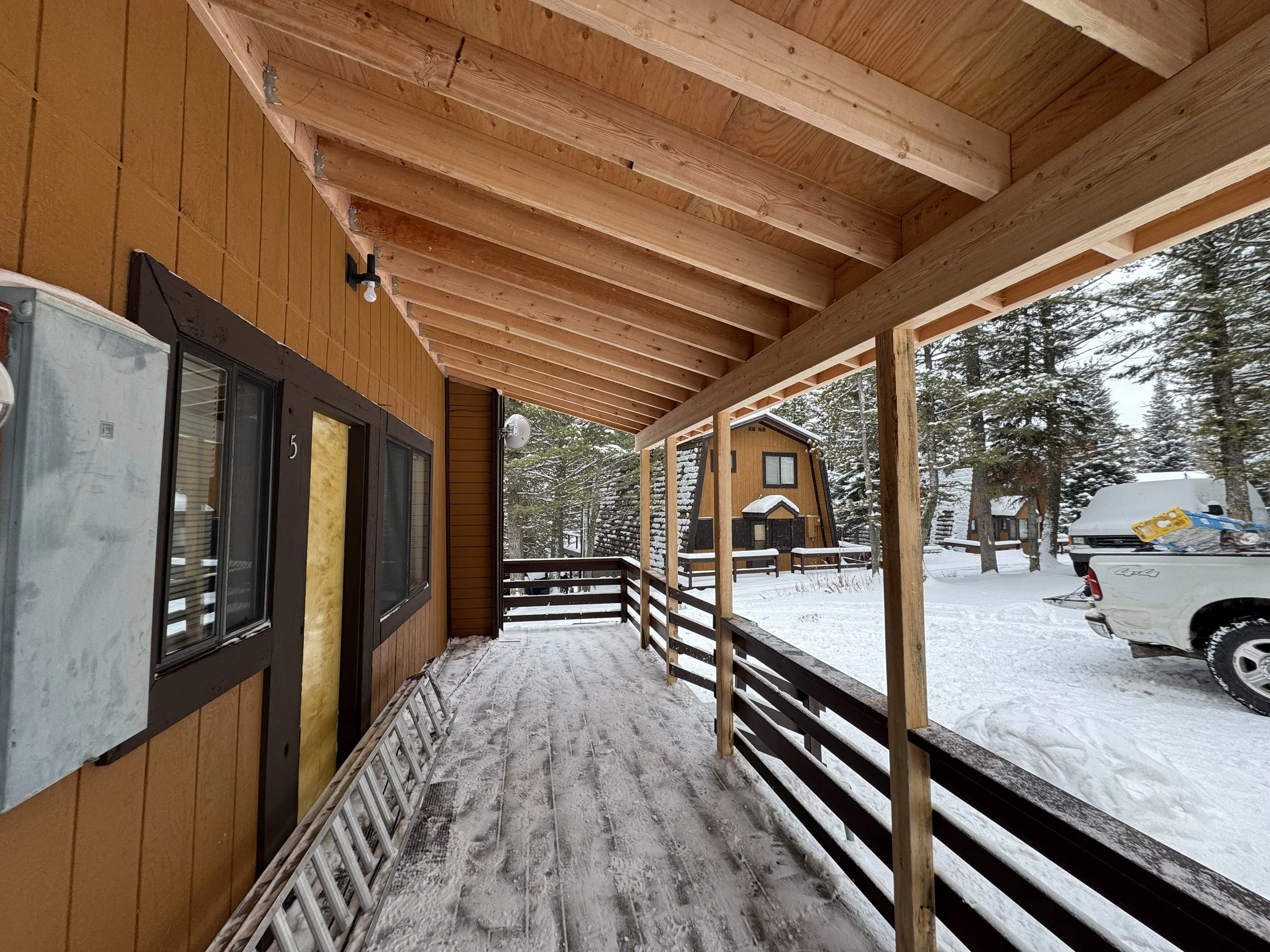 Porch of a wooden house with snow-covered ground and neighboring houses, trees, and a truck in the background.