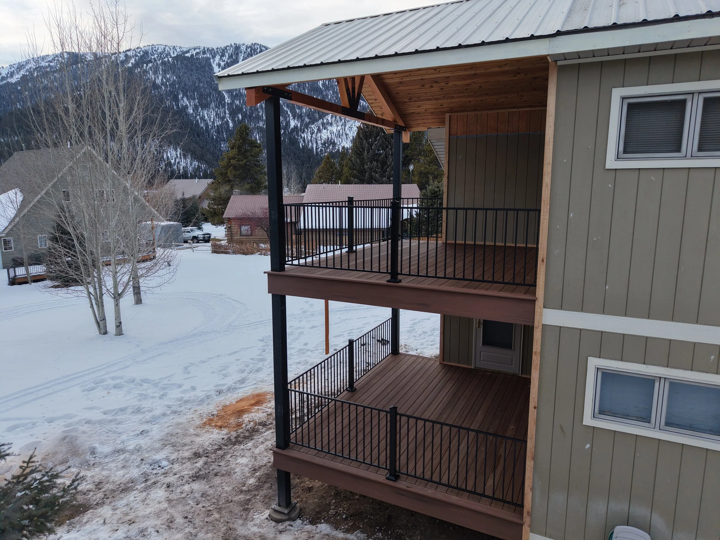 View of a multi-story house balcony with wooden flooring and black metal railings, overlooking snow-covered landscape with trees and mountains.