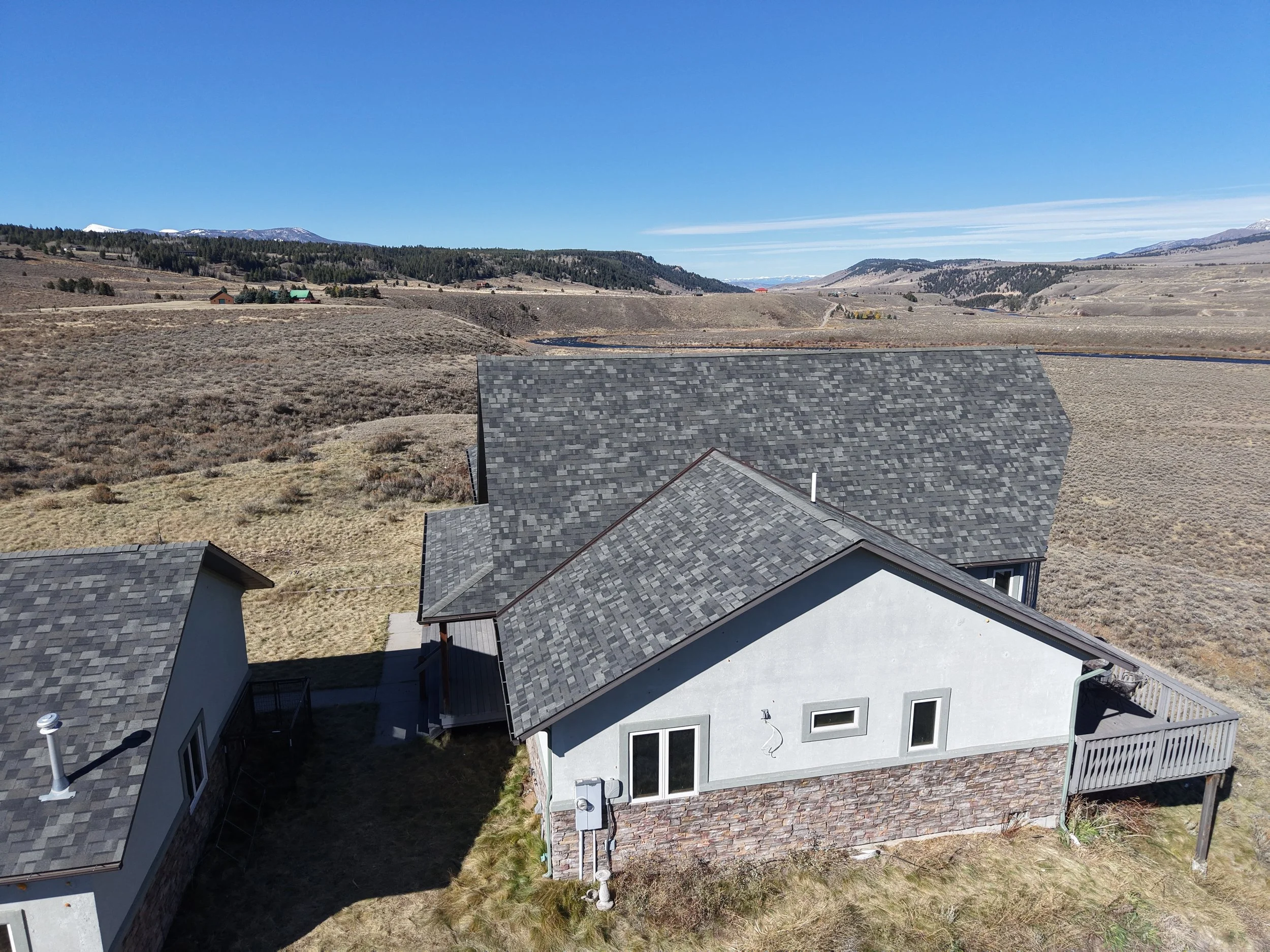 An aerial view of a house with a gray roof and light gray exterior walls, situated in a rural area with open fields and hills in the background under clear blue skies.