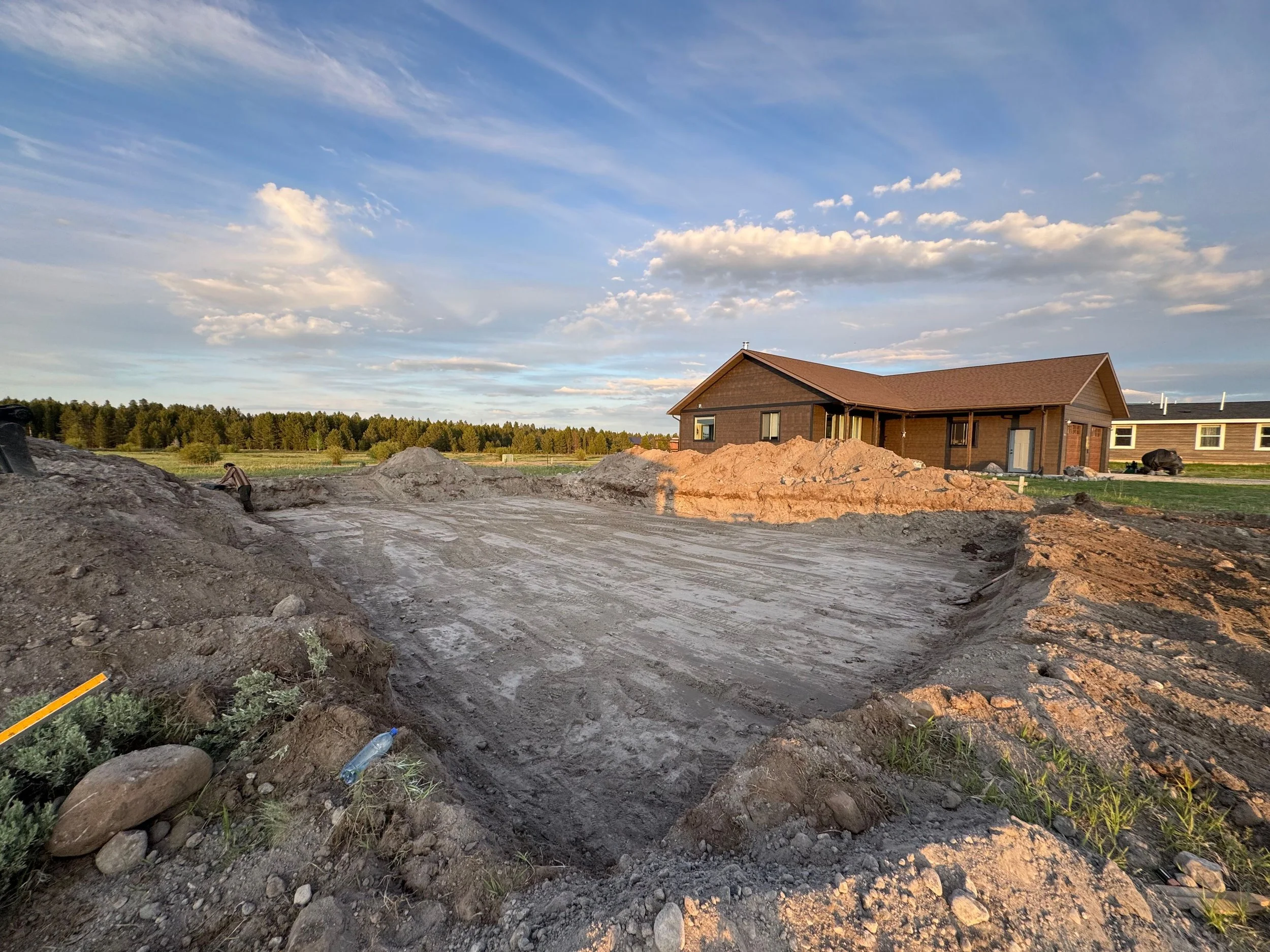 A construction site in front of a house with dirt and excavation marks, with a partly cloudy sky overhead.