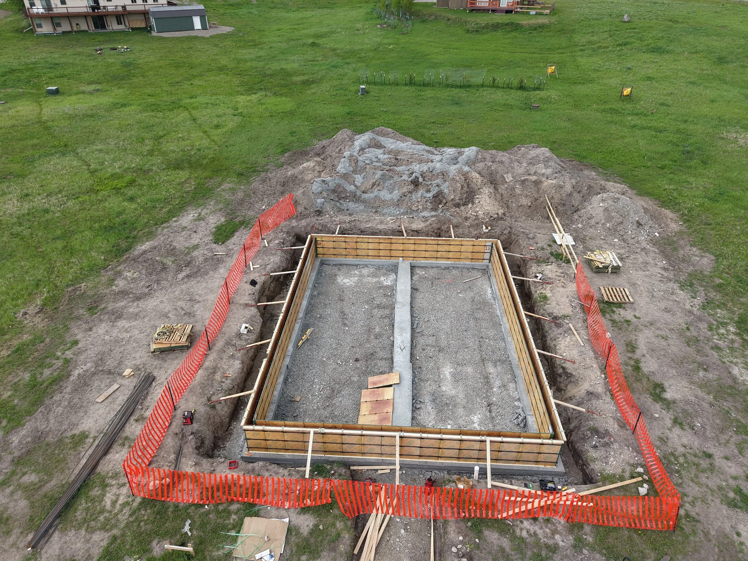 An aerial view of a construction site in a grassy area, showing a partially built foundation with wooden framing, construction tools, and materials surrounded by orange safety barriers.