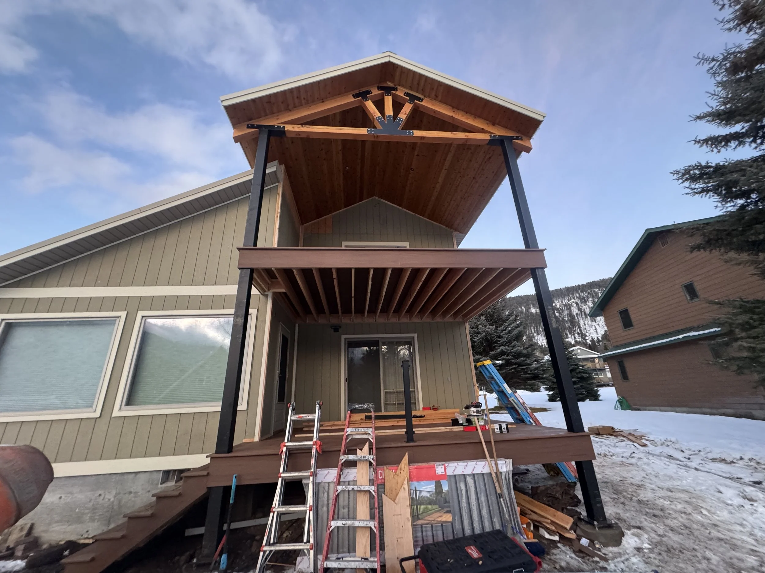 Construction site of a two-story house with a wooden deck and balcony, tools and ladders are visible, snow on the ground, neighboring houses, and a mountain backdrop.