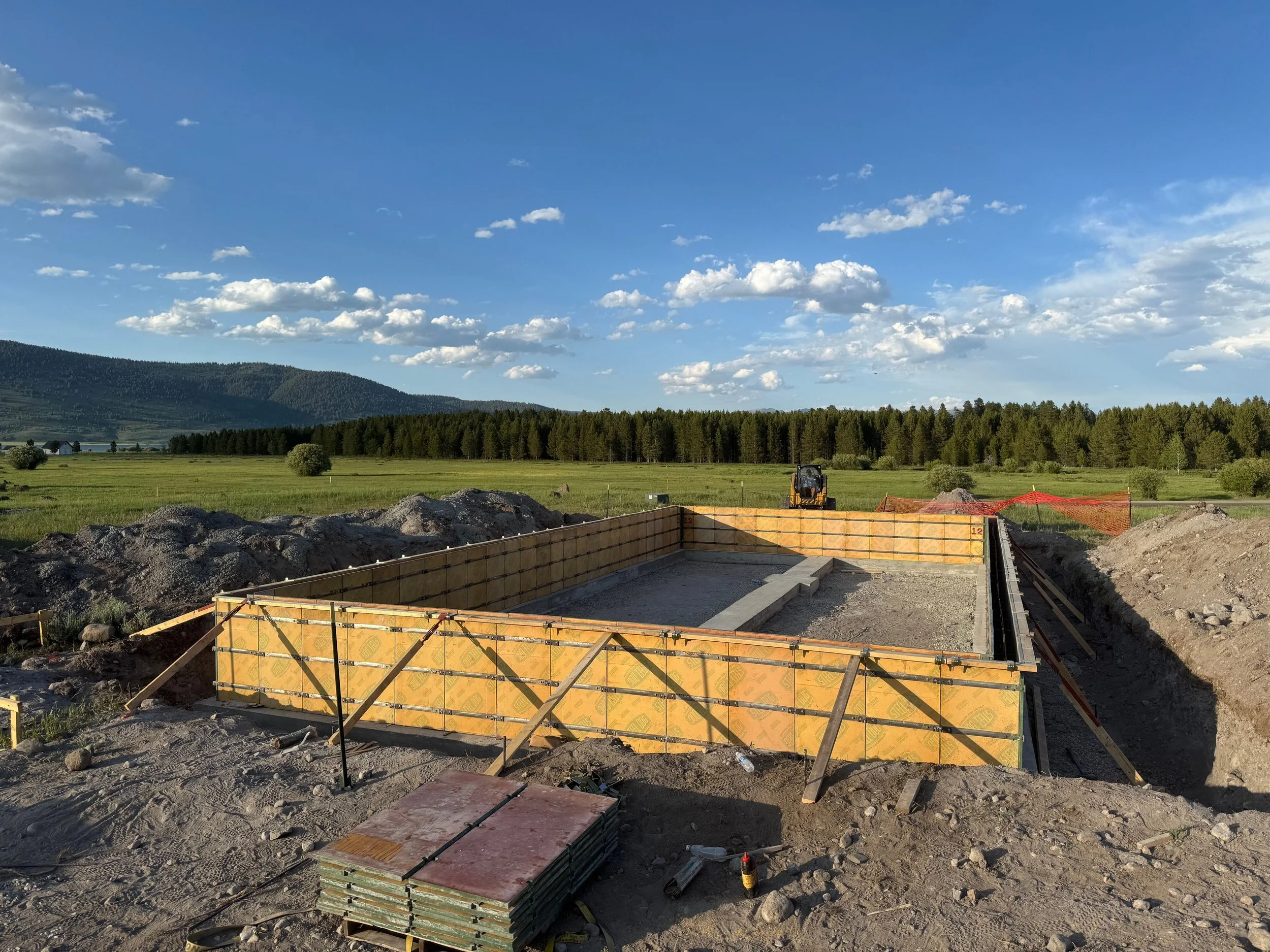 Construction site with a foundation preparing for building, surrounded by open fields, trees, and mountains under a partly cloudy sky.