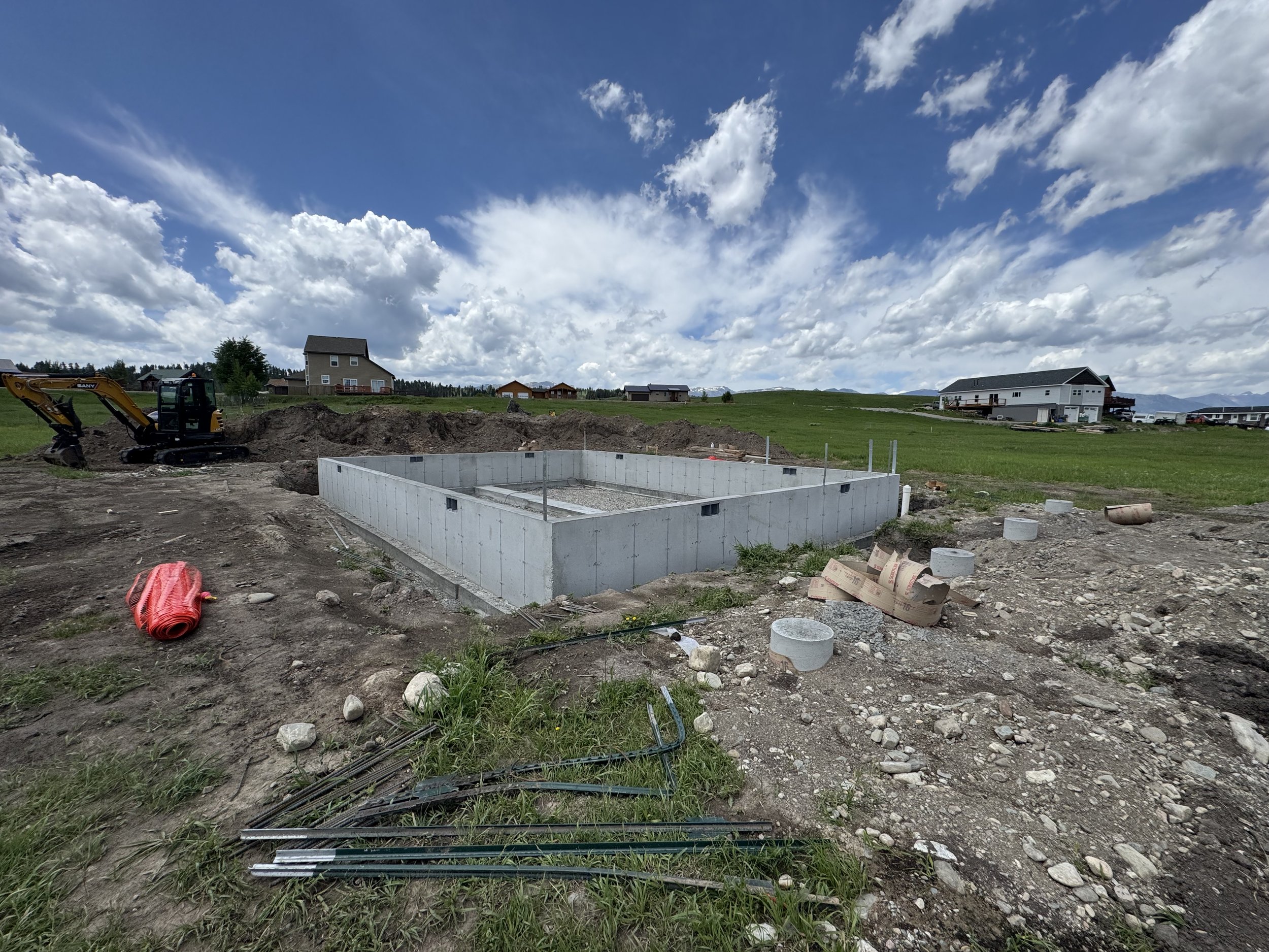 Construction site with a concrete foundation, a small excavator on the left, scattered construction materials, and houses in the background under a partly cloudy sky.