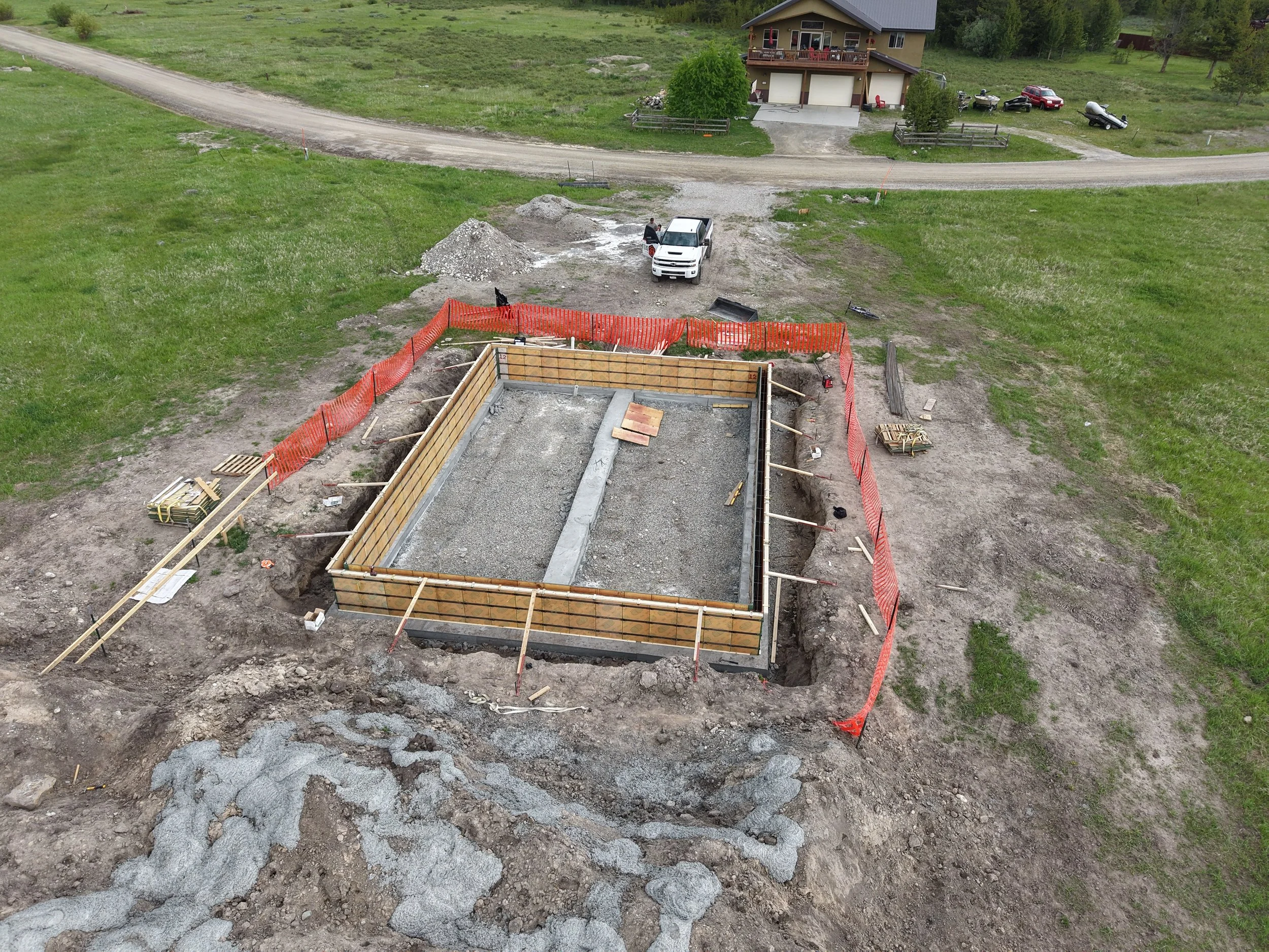 A construction site with a foundation being built for a house, surrounded by orange safety fencing. Construction materials and tools are scattered around. There is a gravel area and a dirt path leading to a nearby house, with vehicles parked in the b