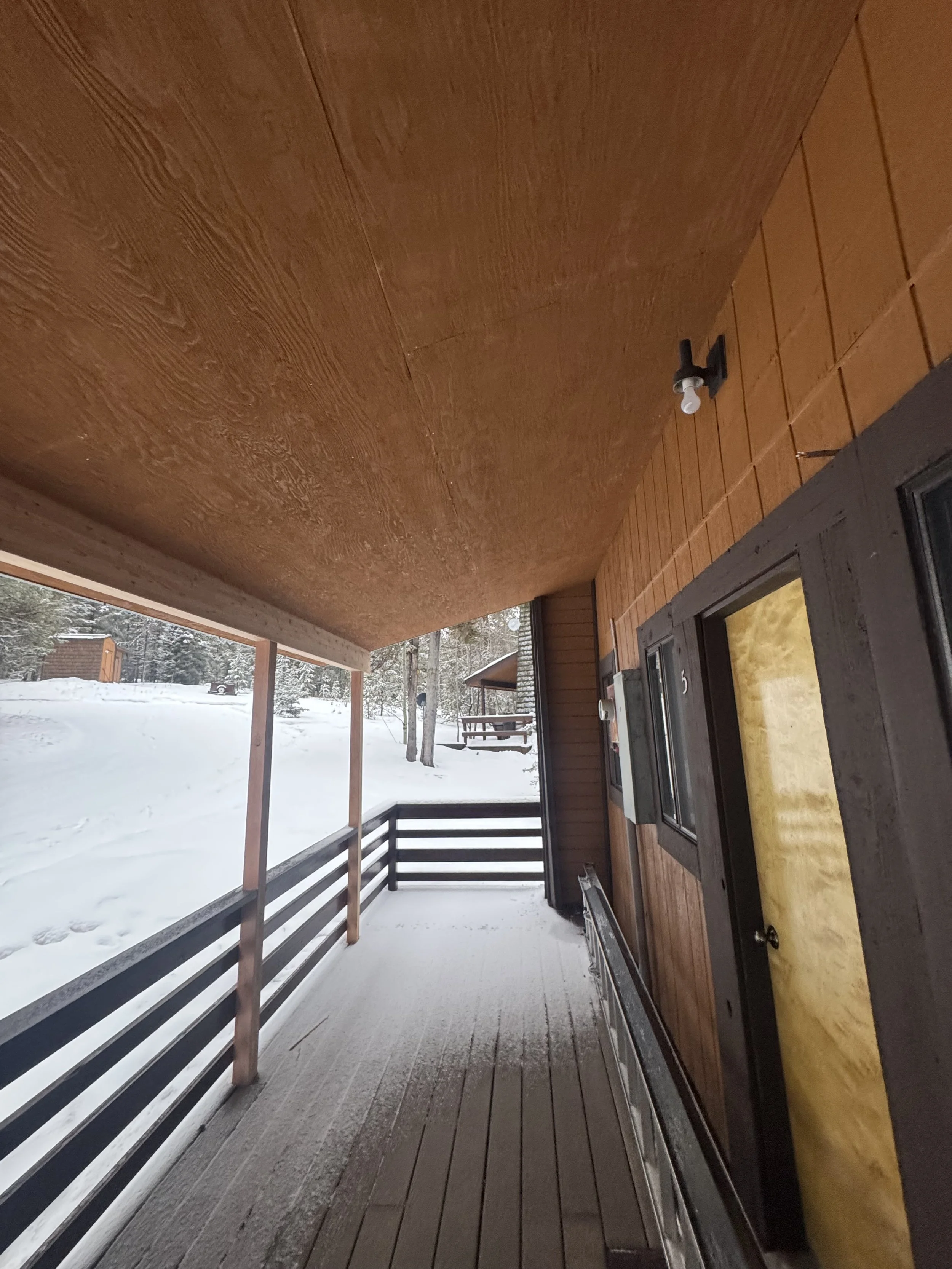 Snow-covered porch of a cabin with wooden railing, next to a snowy landscape with trees and other cabins in the distance.