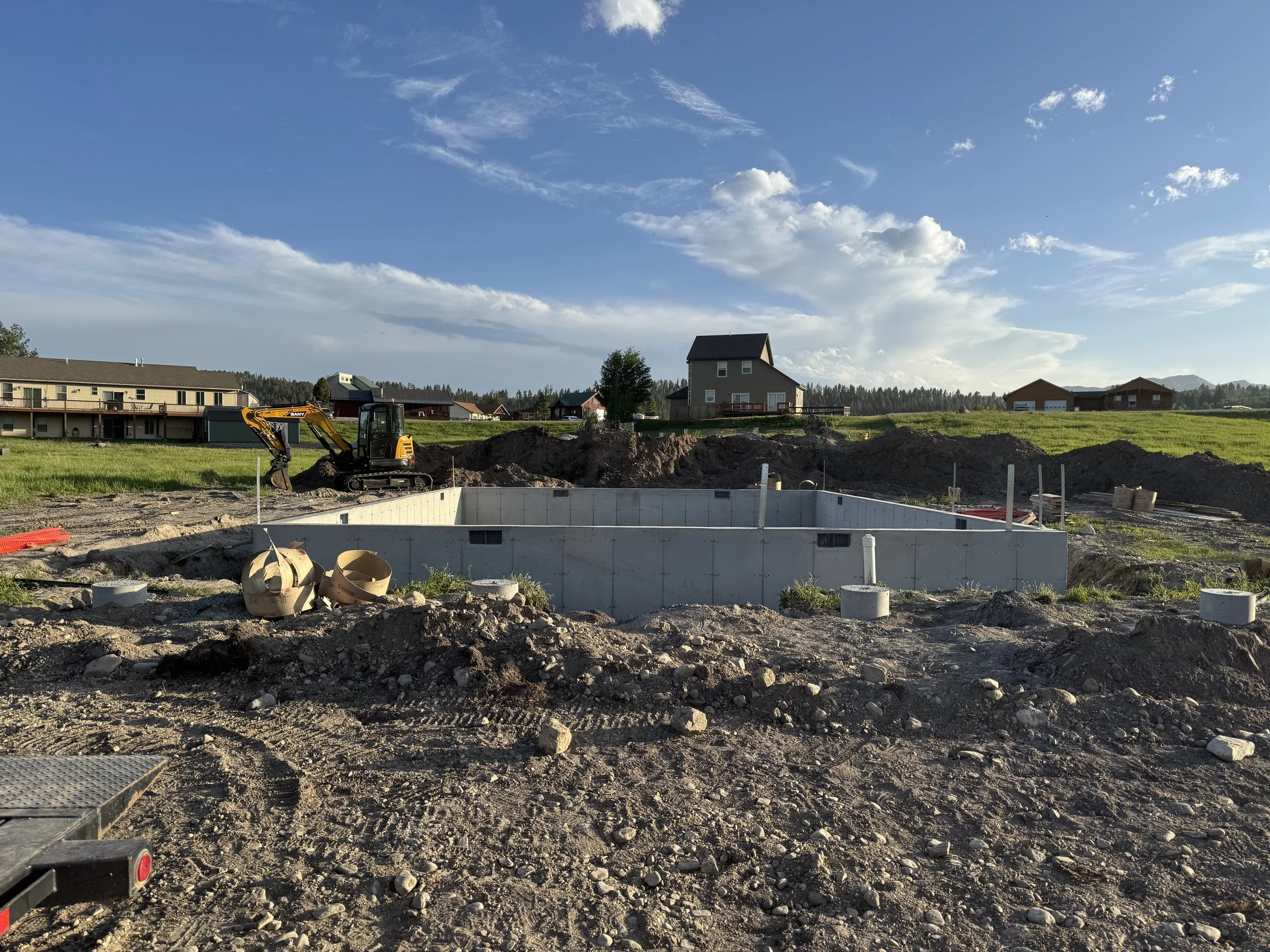 Construction site with a concrete foundation and a small yellow excavator, surrounded by dirt and construction materials, under a partly cloudy sky.